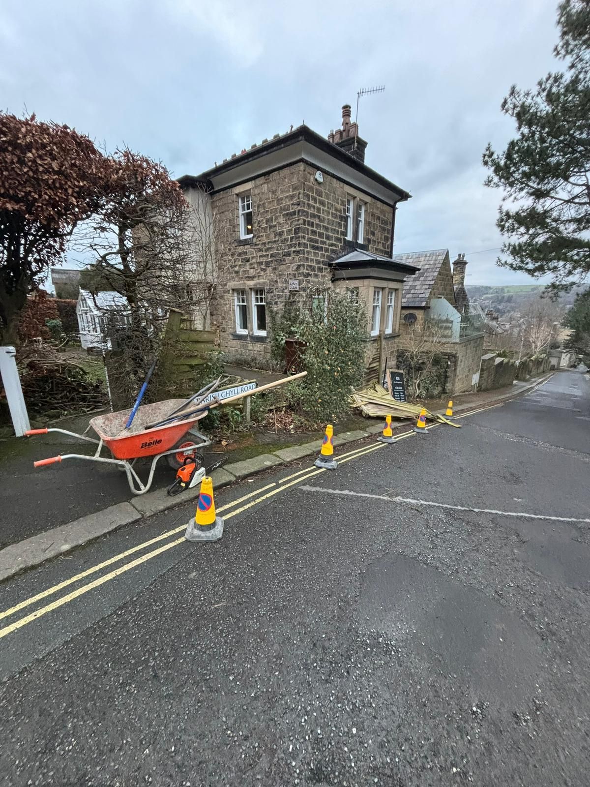 Roadside stone house with cones, orange barriers, and a wheelbarrow blocking a wet lane