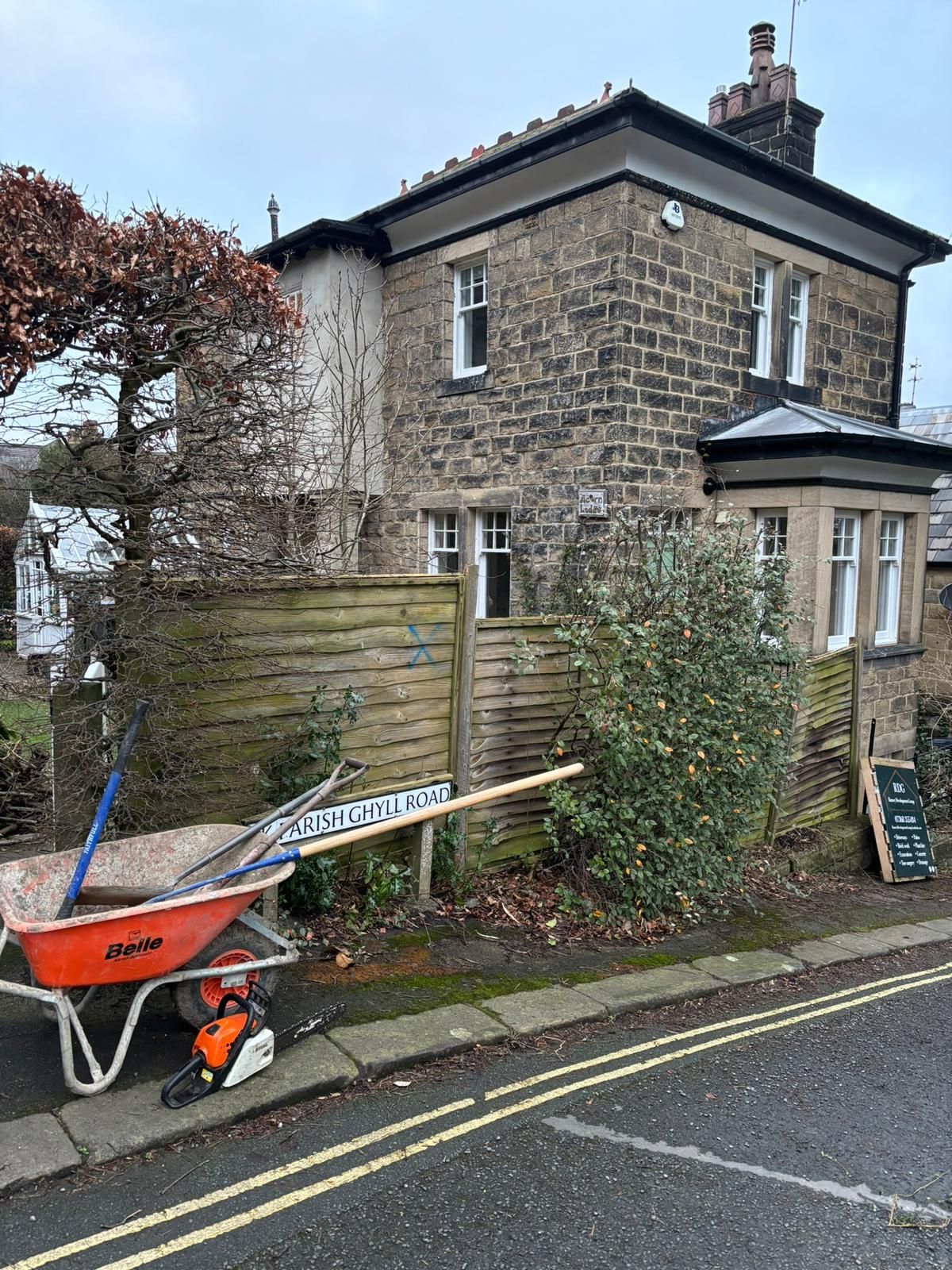 Stone house beside road with orange wheelbarrow and wooden fence in front garden
