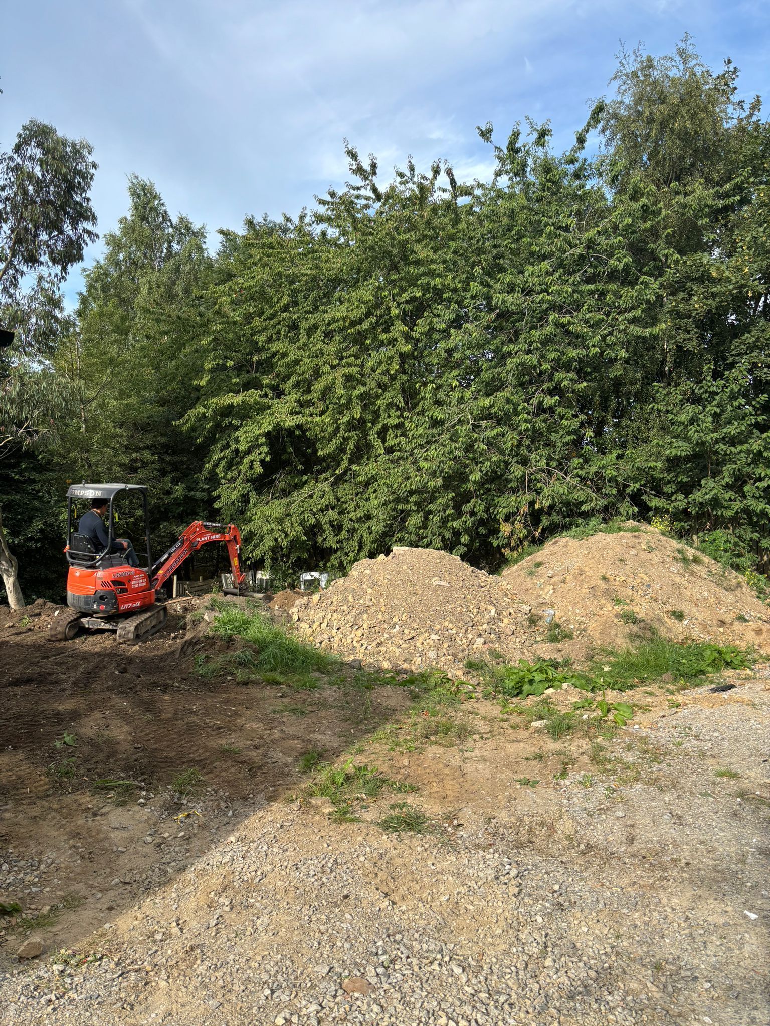 Small red excavator beside gravel piles and dense green bushes under a blue sky.