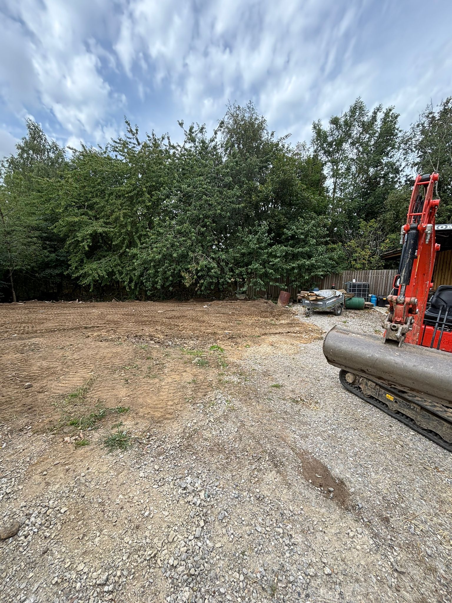 Gravel lot beside dense trees, with construction equipment and a red excavator arm on the right.