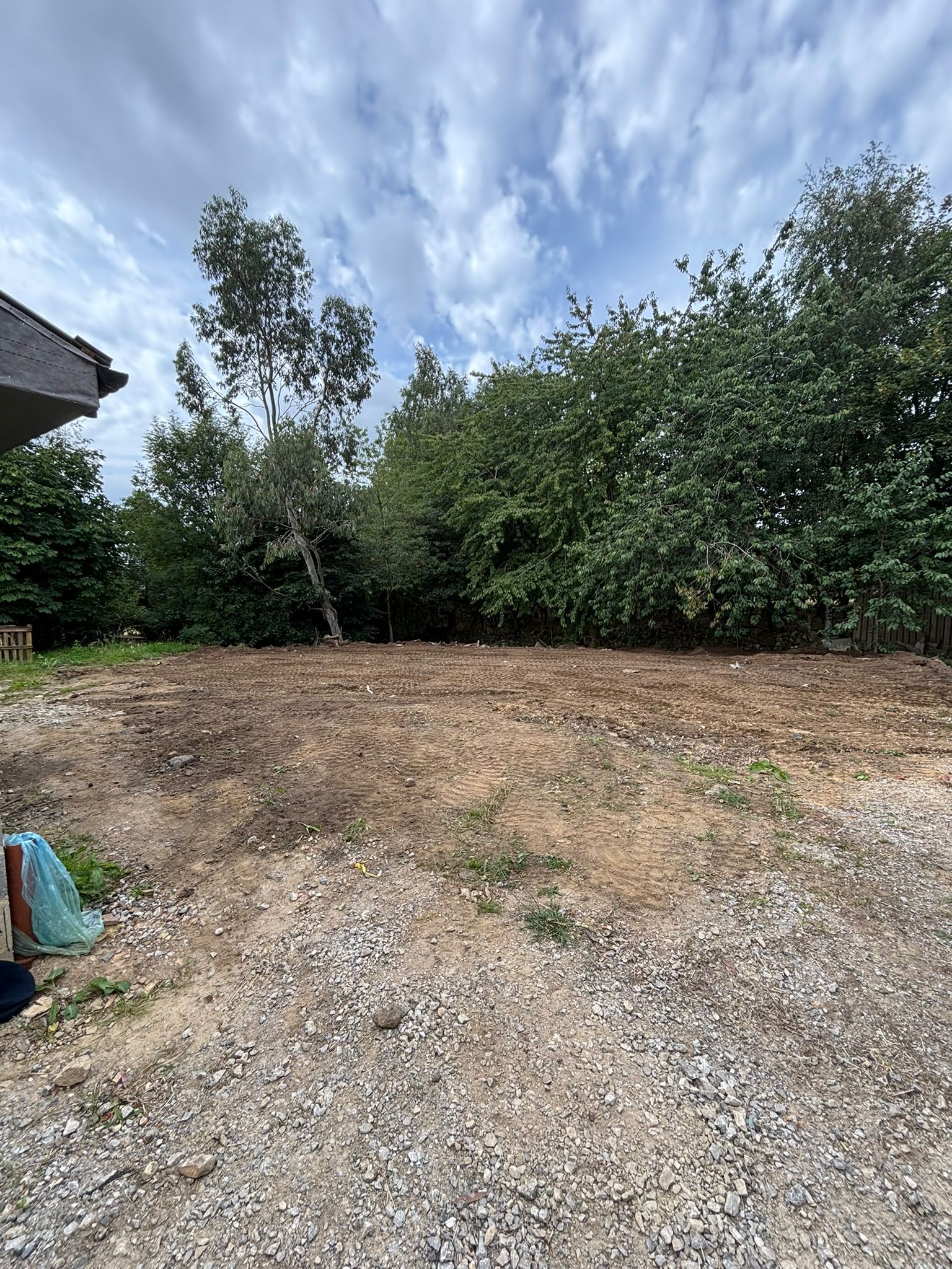 Gravel driveway leading to a tree-lined yard under a cloudy sky, with a partial house roof at left