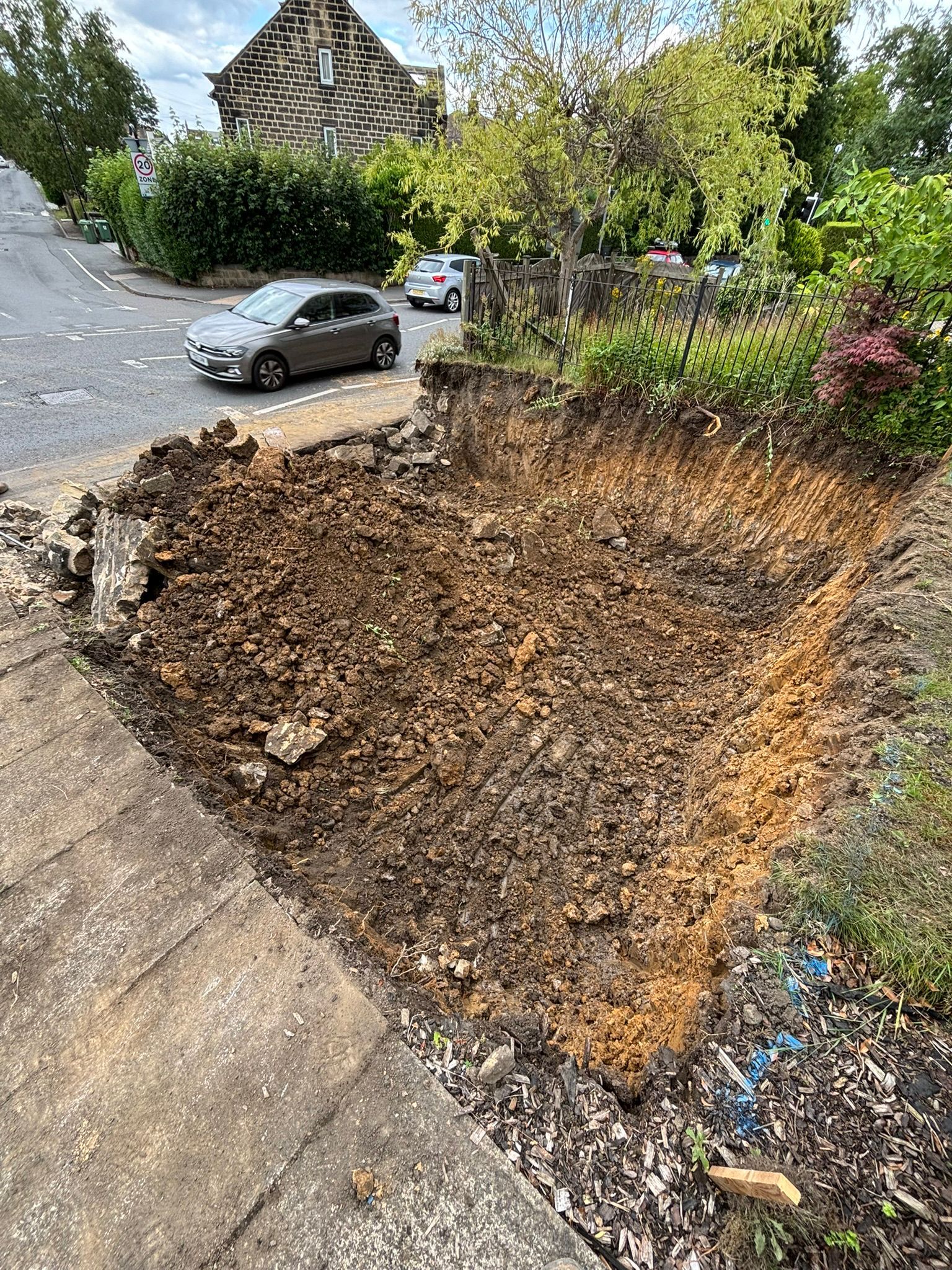 Large eroded roadside pit beside a driveway, with parked cars and trees in the background.