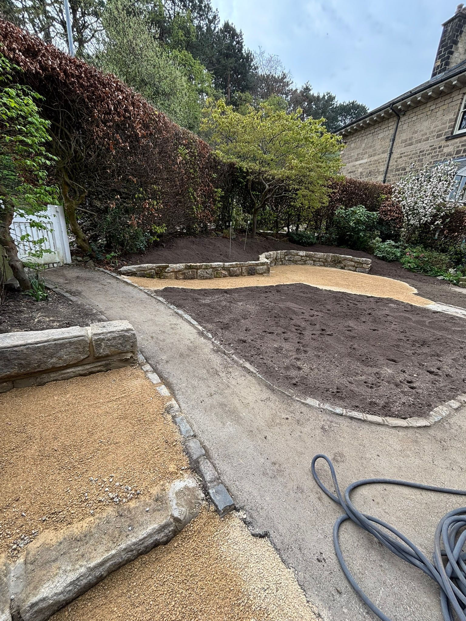 Garden path with gravel, soil beds, stone steps, and a coiled hose beside a stone house