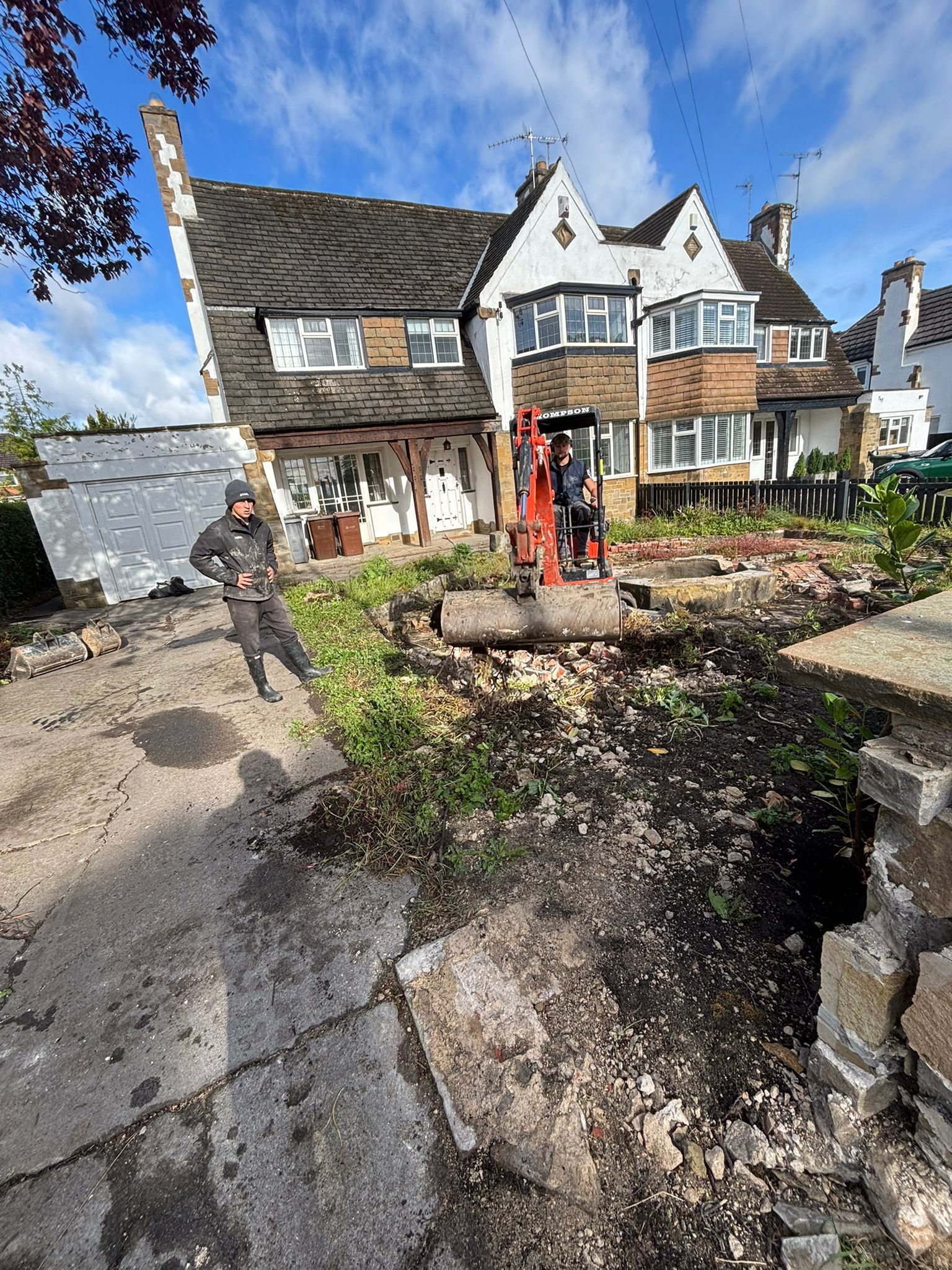 Dirt driveway beside a stone cottage with a red excavator and a partially dug garden under a blue sky