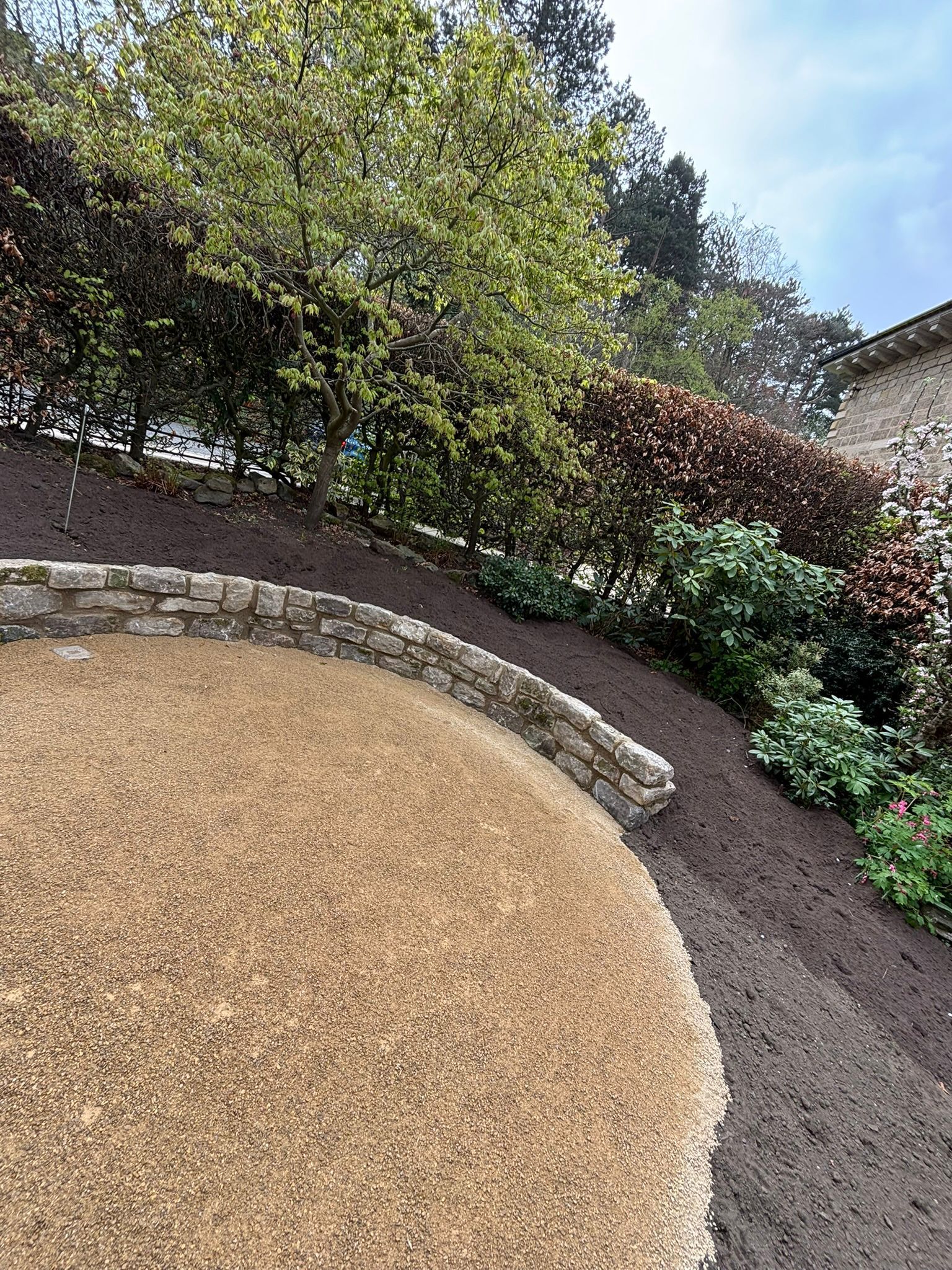Curved gravel path beside a landscaped garden with trees, shrubs, and stone edging