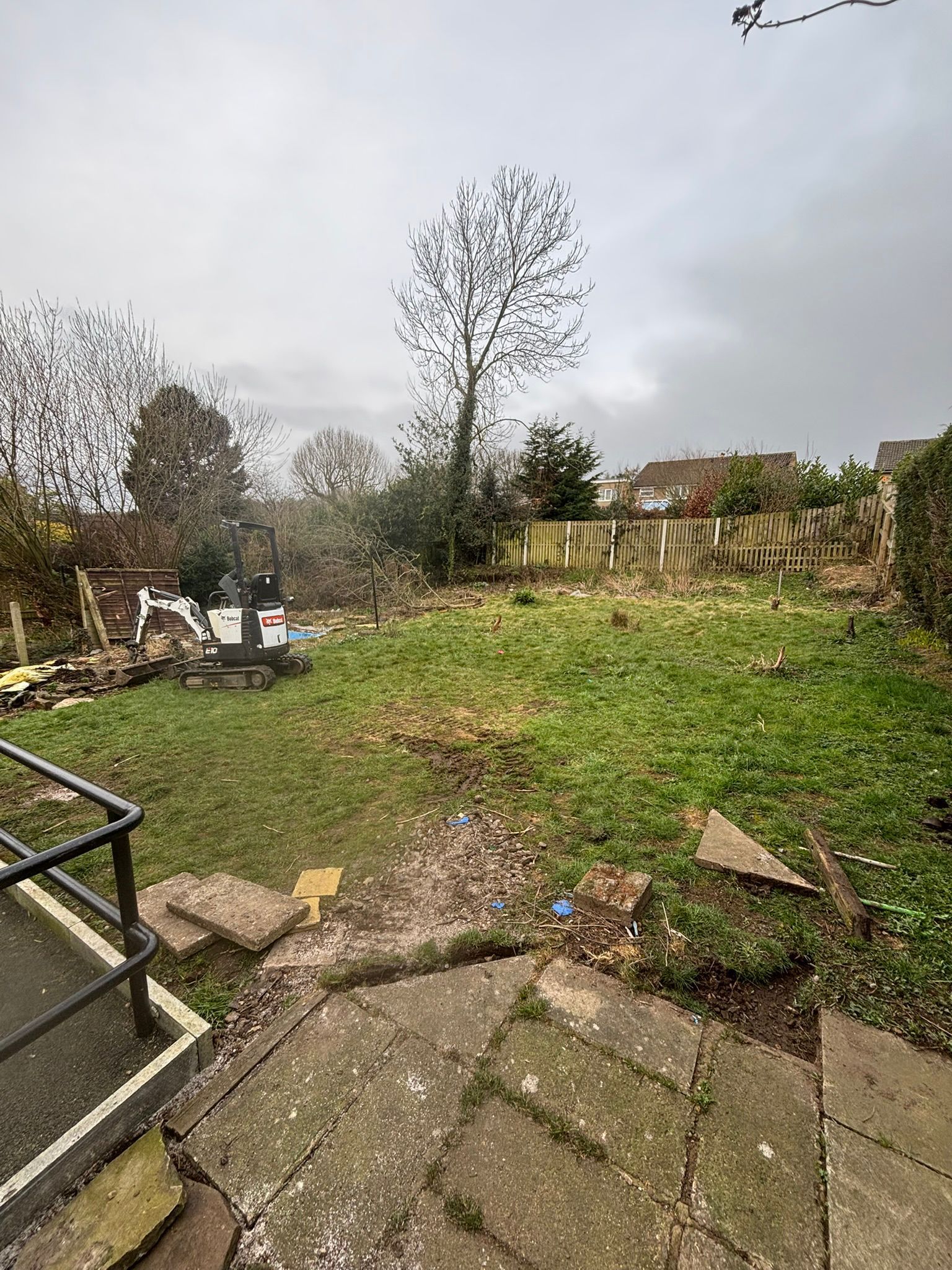 Grassy backyard with stone patio, metal railing, bare trees, and a cloudy sky.