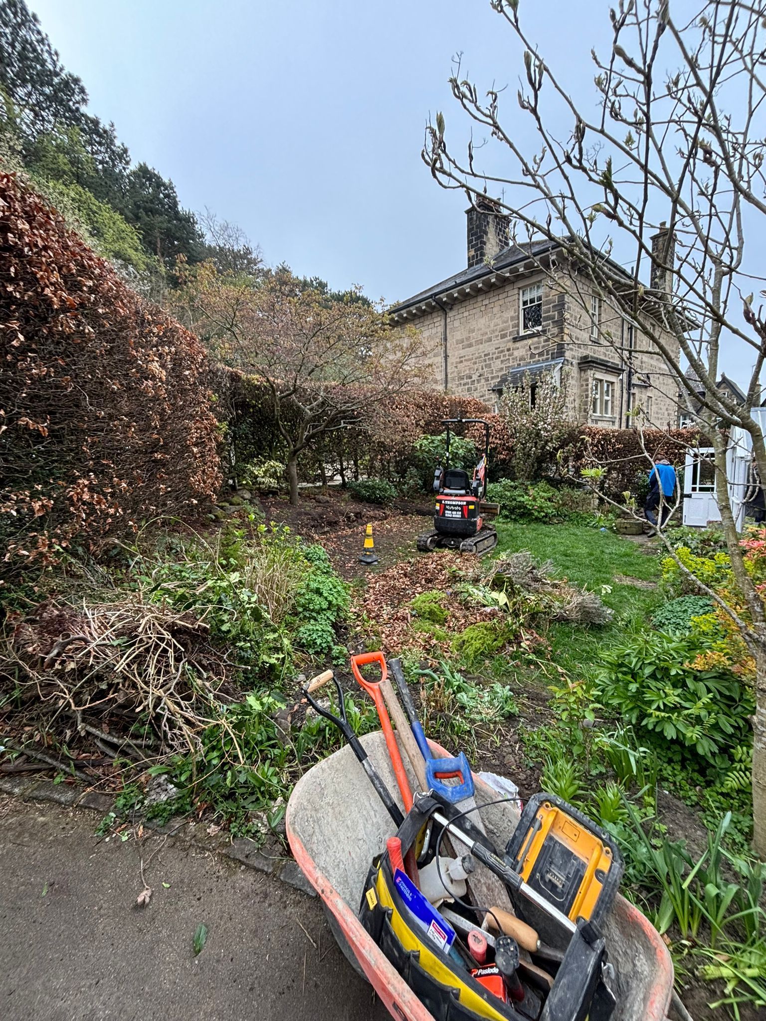 Wheelbarrow on a sloped garden path with tools, shrubs, and a house in the background.