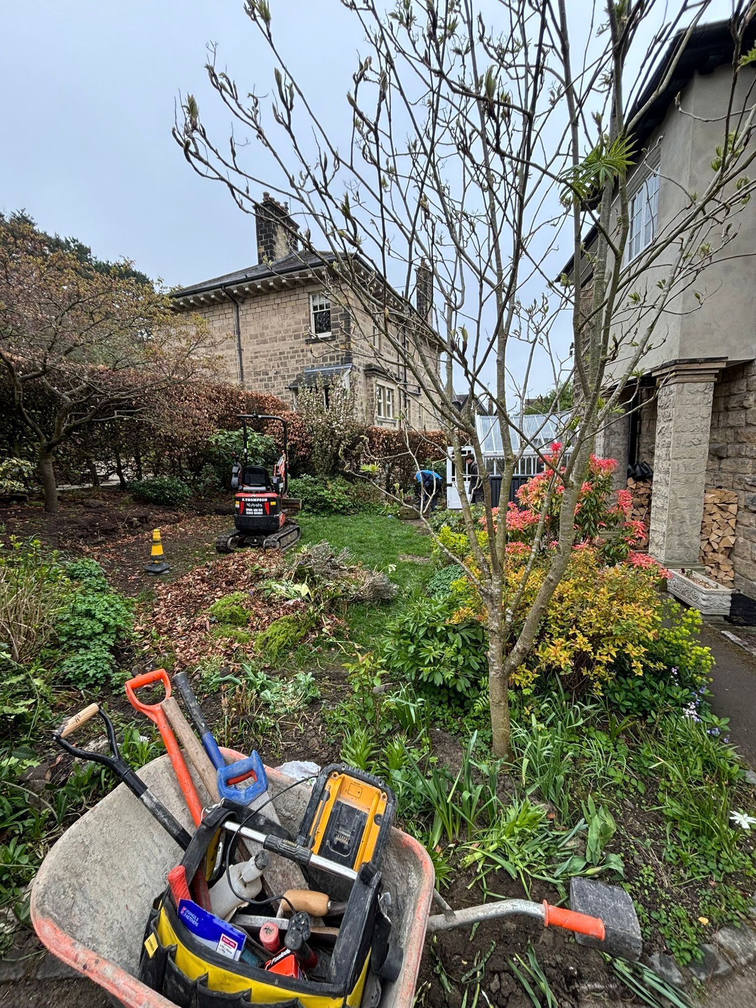 Garden path with wheelbarrow and tools beside a house, bordered by plants and flowers.