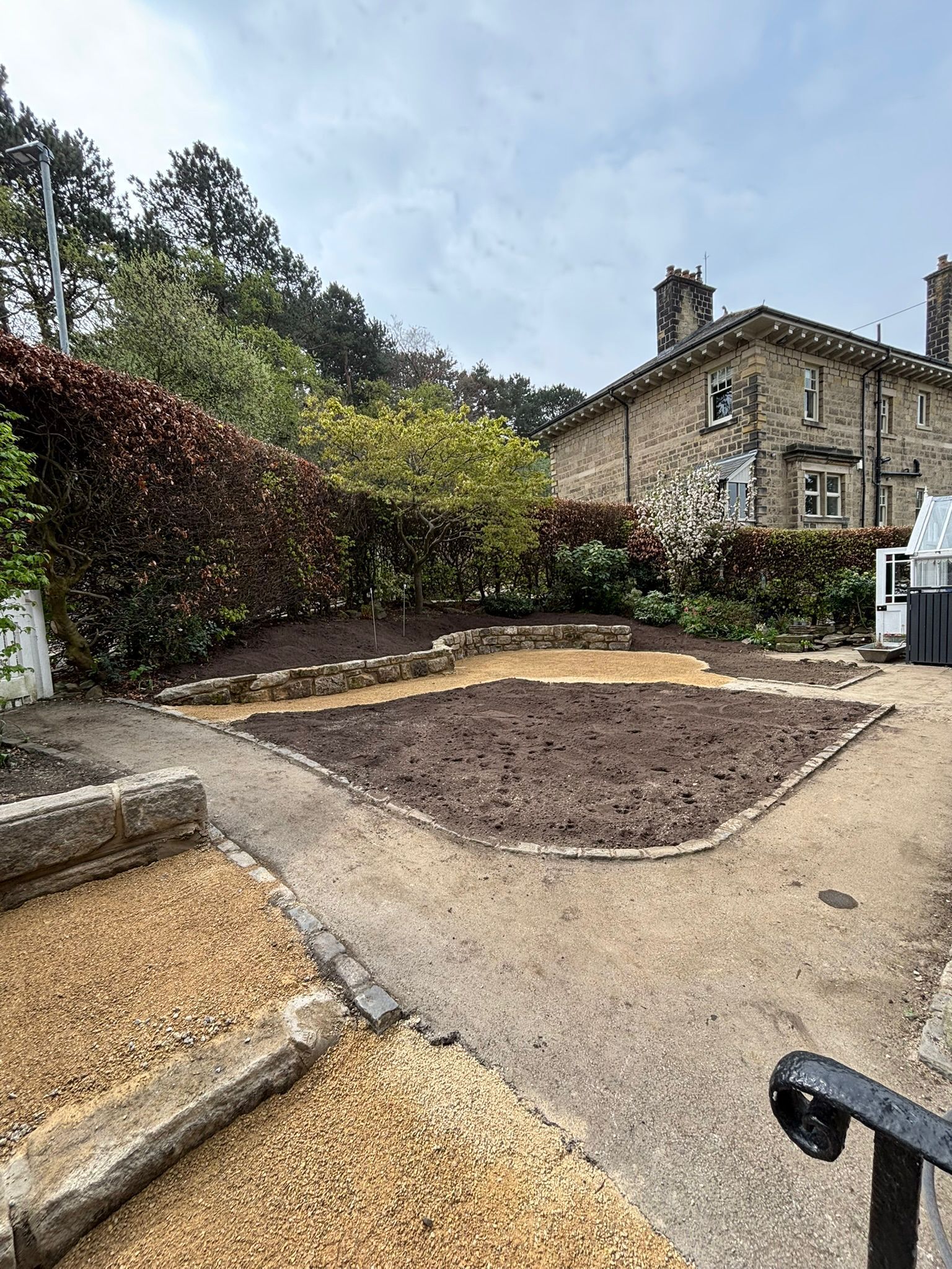 Garden path with fresh soil beds beside a stone house and hedges under a cloudy sky