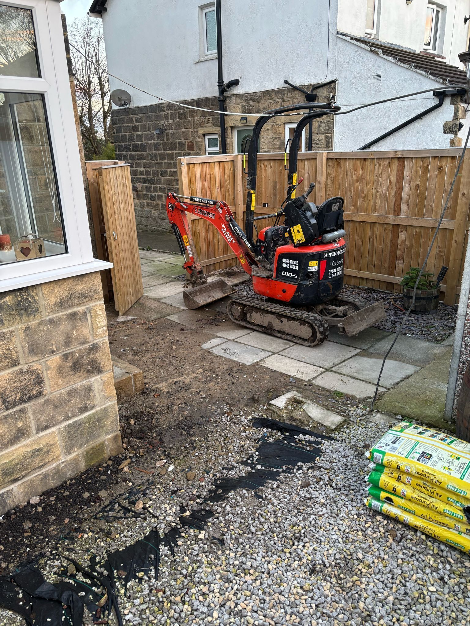 Small red excavator in a backyard beside a fence and gravel, with paving stones and stacked bags.