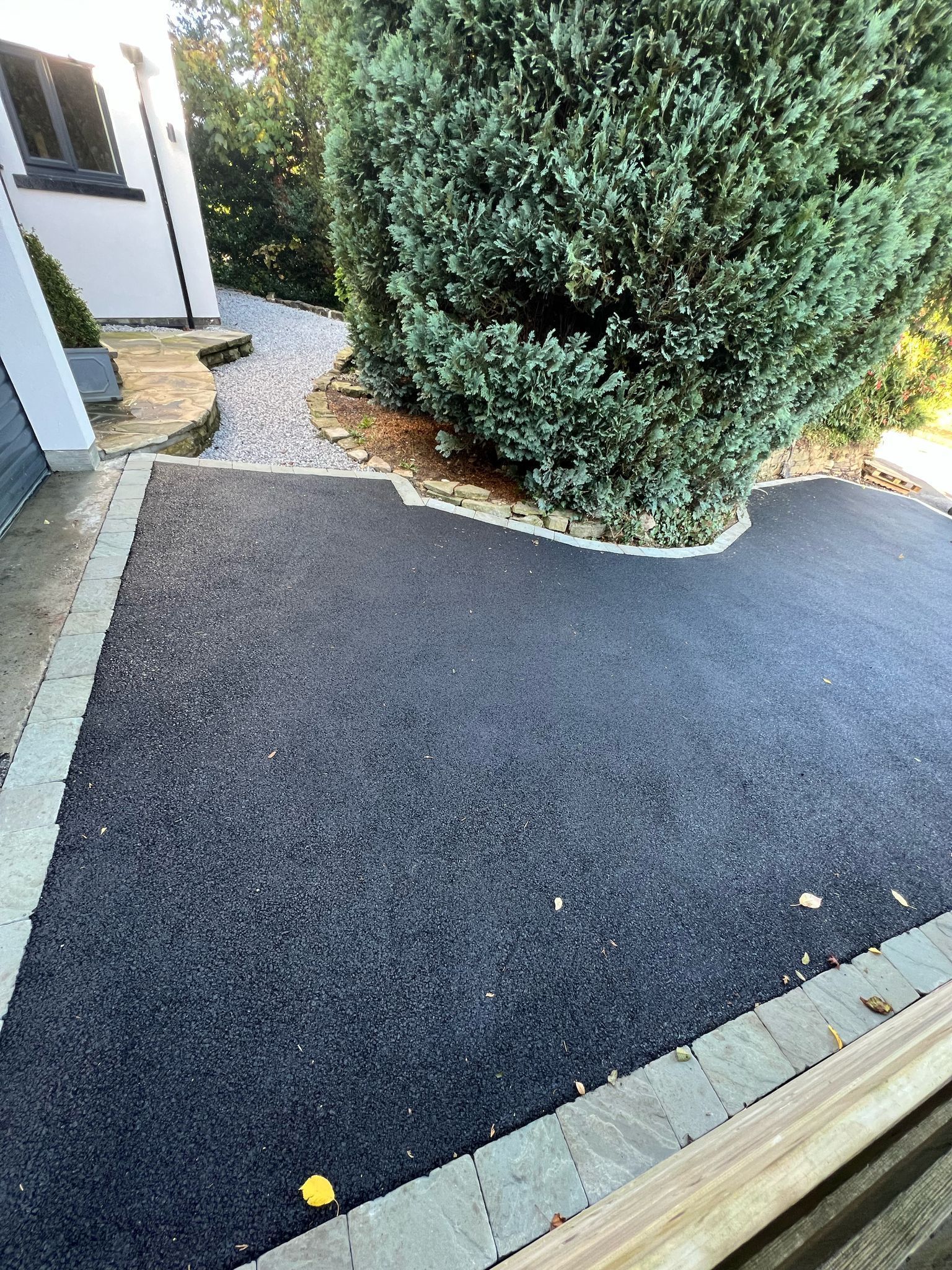 Driveway and sidewalk beside a large bush, with a dark paved surface and light stone edging.