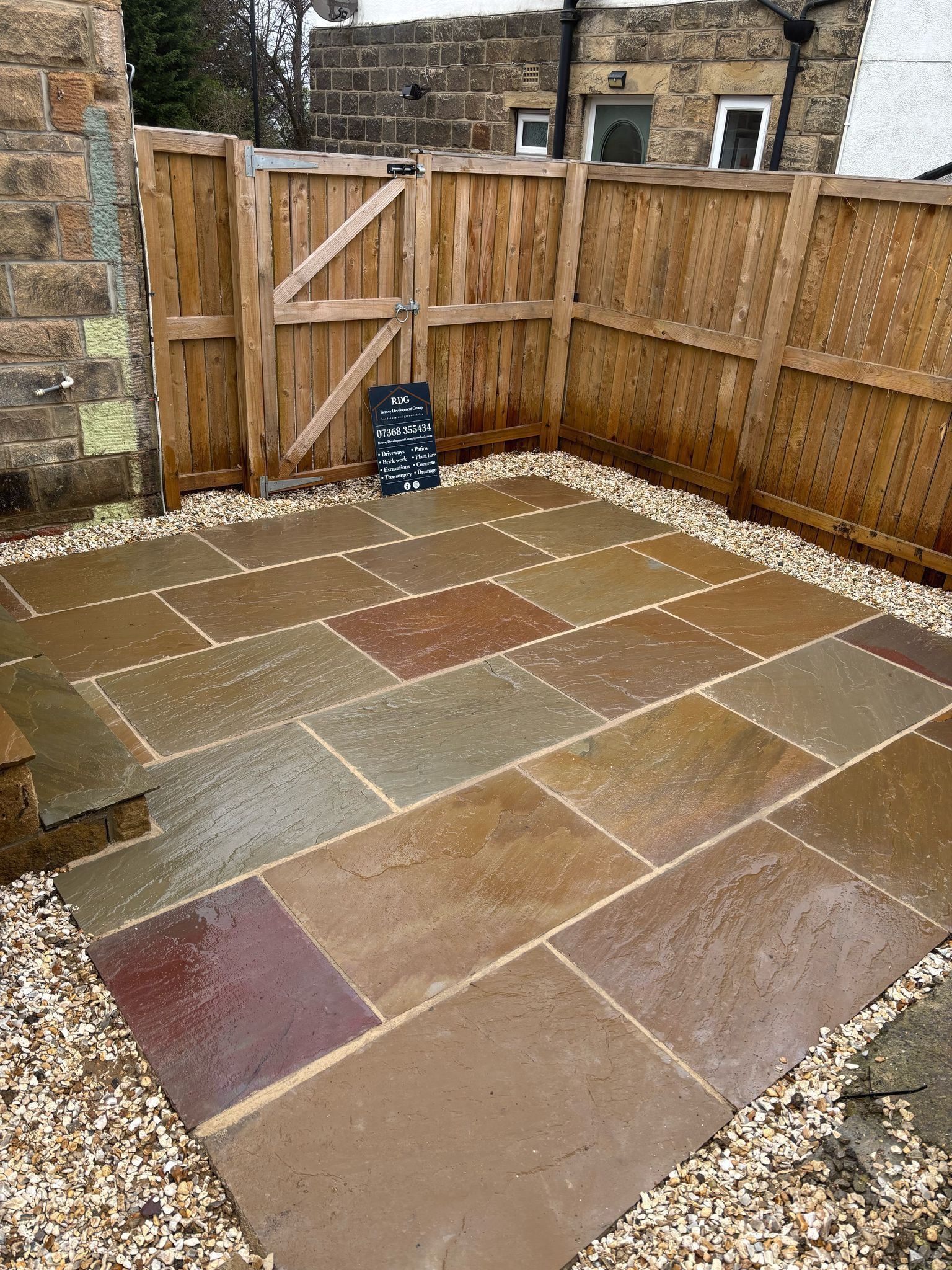 Paved backyard patio with multi-colored stone slabs, gravel edging, and wooden fencing and gate.