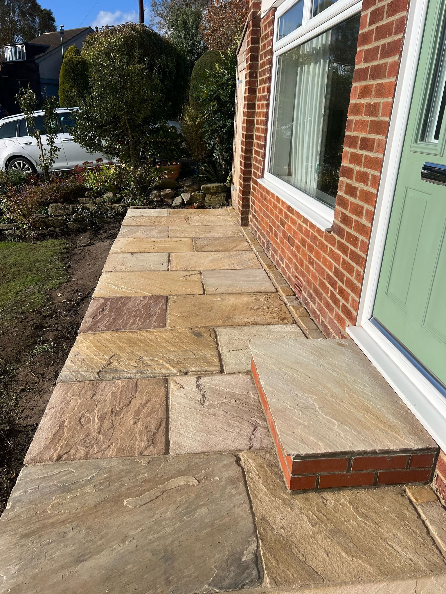 Front walkway of tan stone pavers beside a brick house with a green door and window.