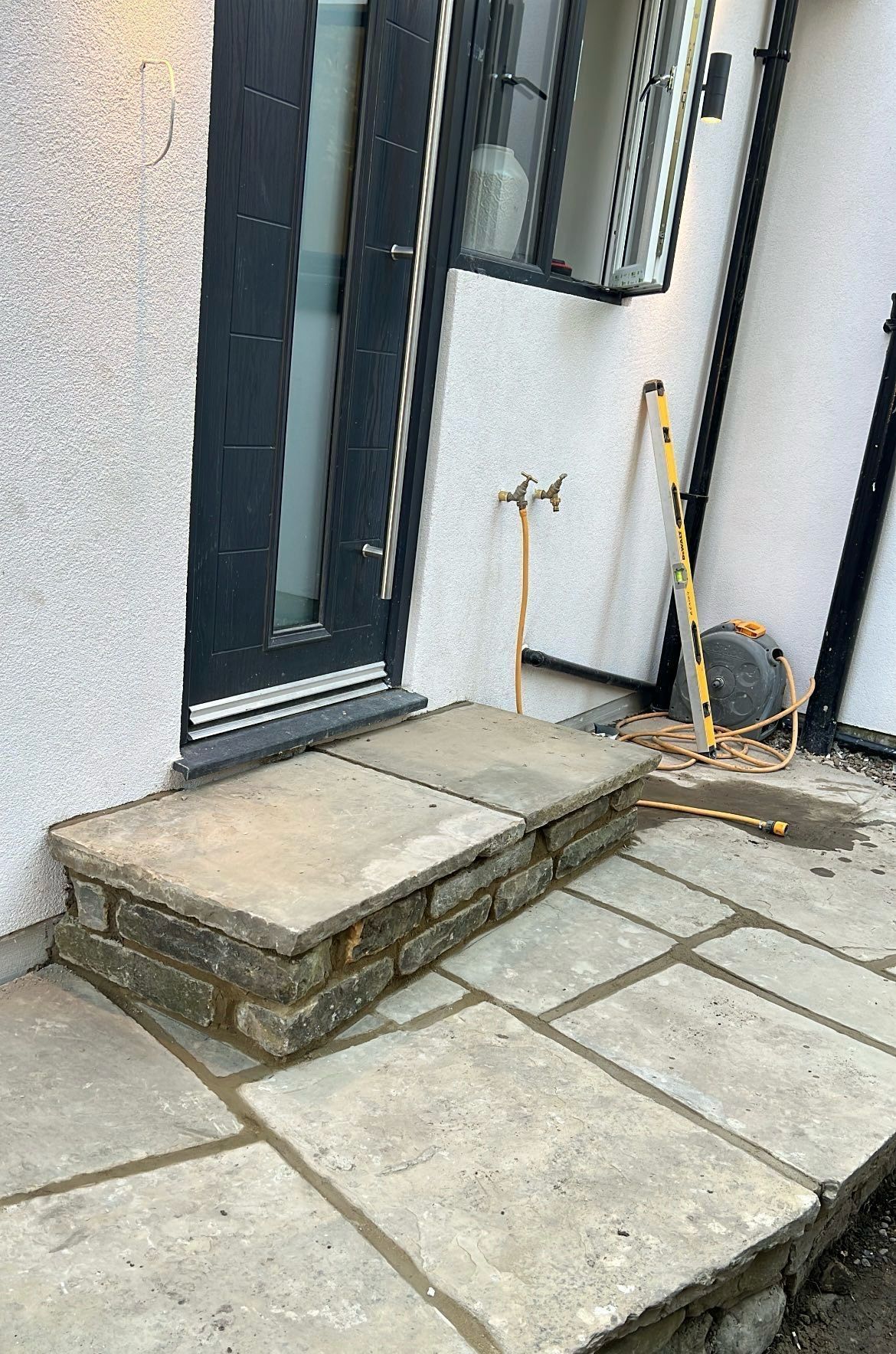 Front entrance with a concrete step and patio slabs beside a dark door and cleaning tools on the right.