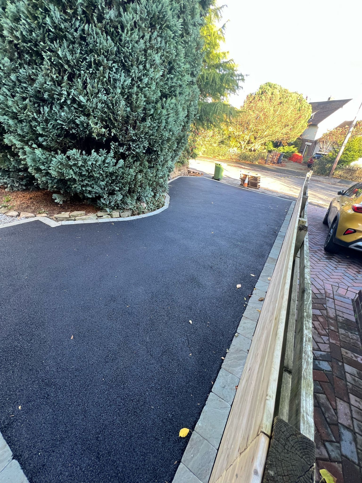 Freshly paved driveway beside a large evergreen tree, with a yellow car parked along a brick curb.