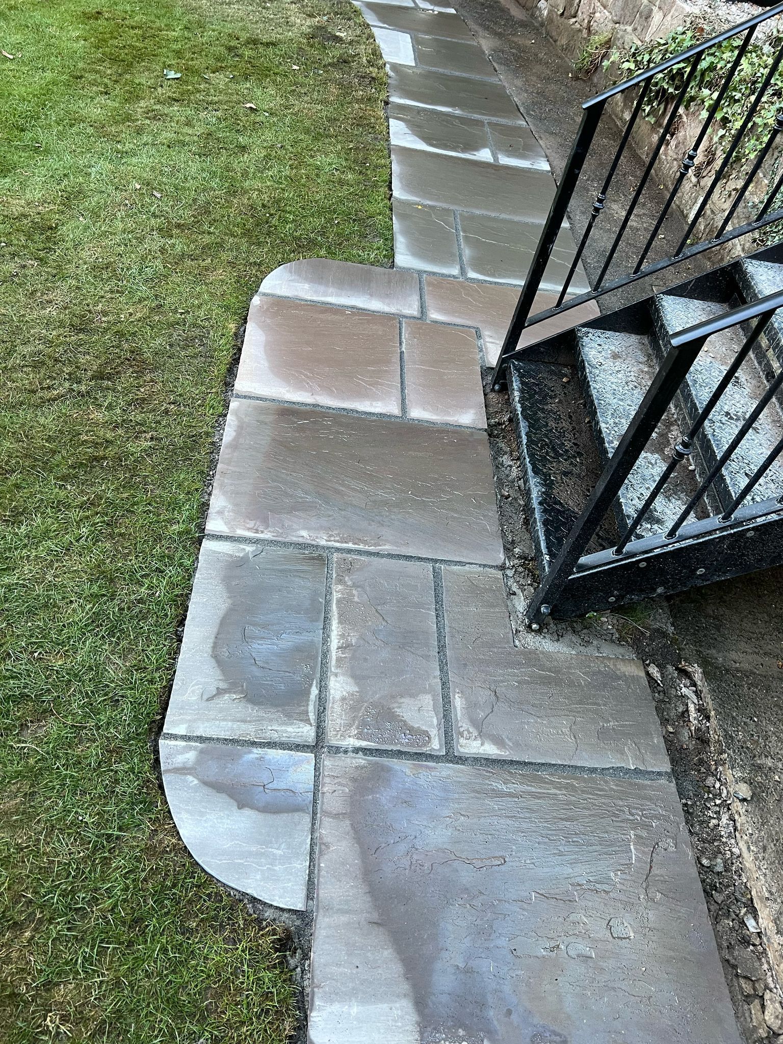 Stone walkway beside grass and a black metal gate, with a curved paver edge.