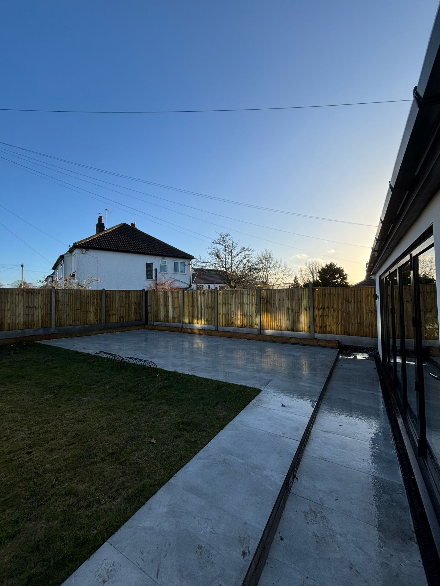 Backyard patio at dusk with concrete path, grass, wooden fence, and a house in the distance
