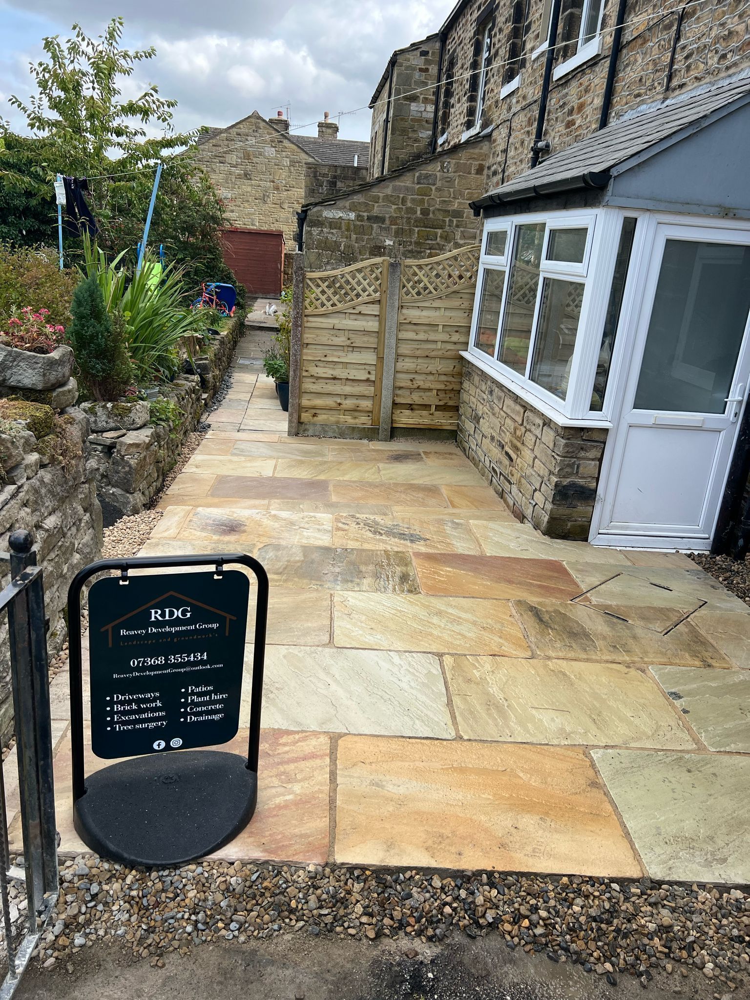 Stone patio beside a stone house with white bay windows, a black A-board sign, and a wooden fence.