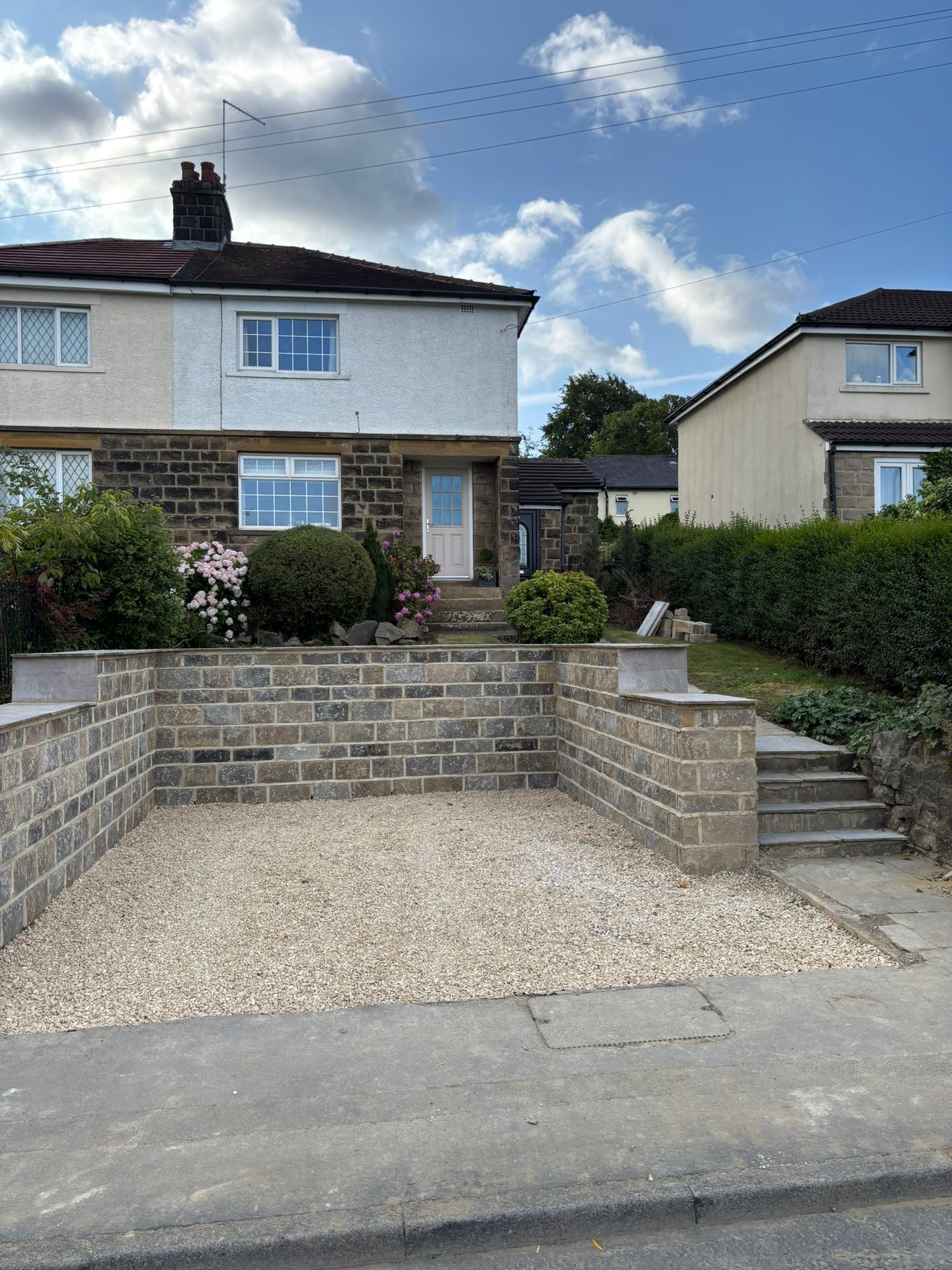 Semi-detached house with front gravel driveway, stone steps, hedges, and blue sky.