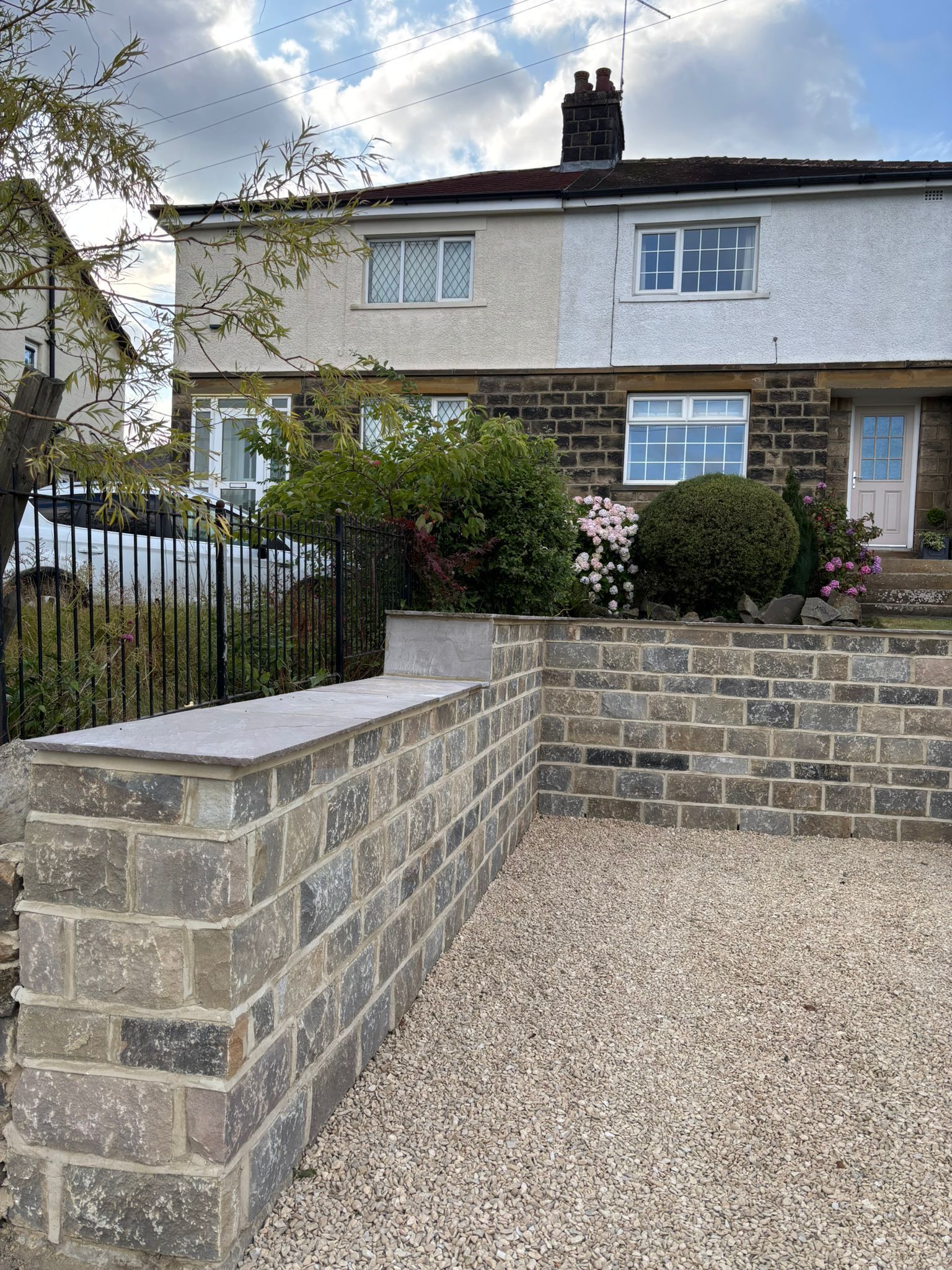 Stone front wall and gravel path leading to a white-and-gray house with garden shrubs and a black gate