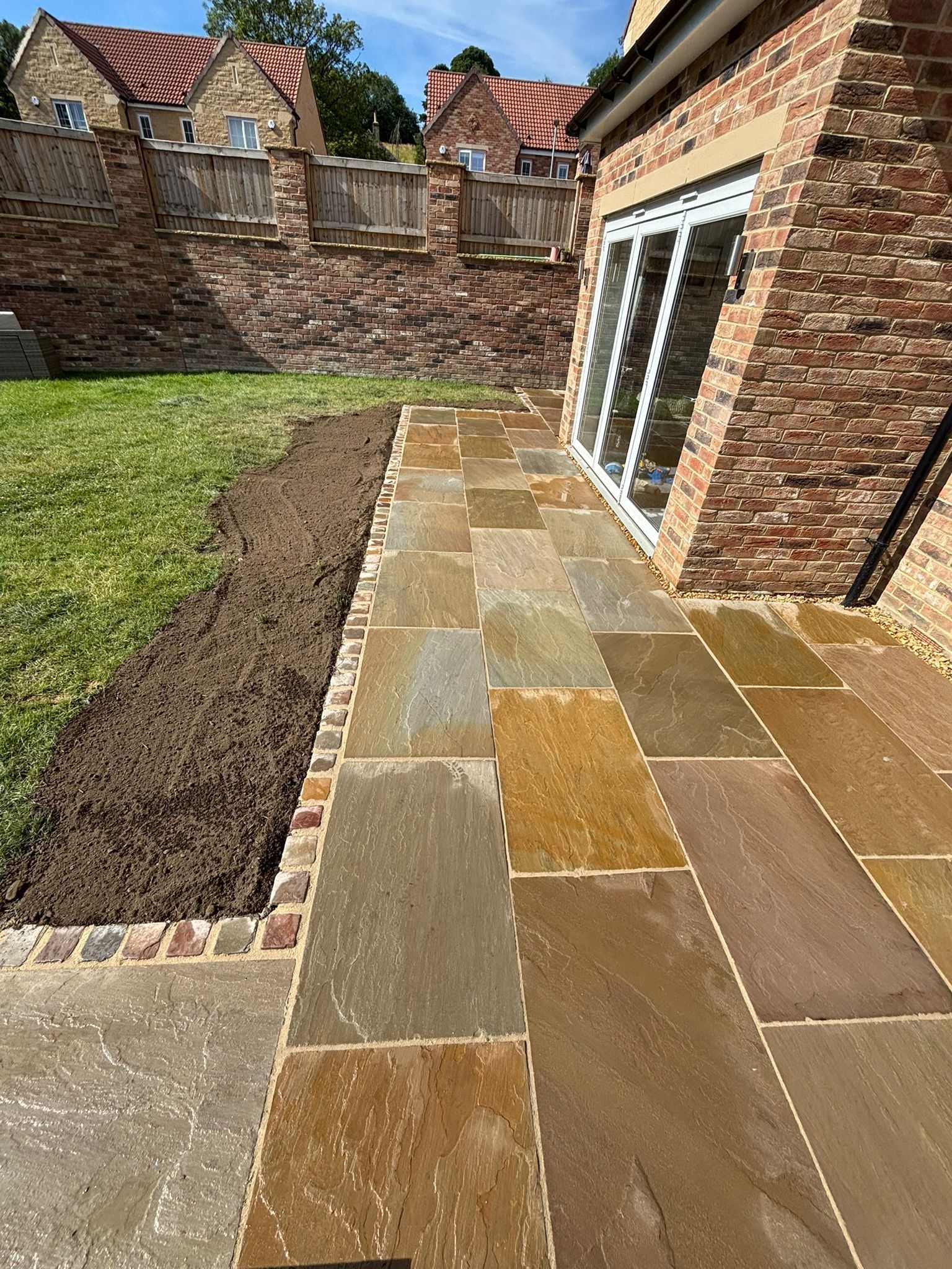 Stone patio beside a brick house, bordered by lawn and a brick wall in a sunny backyard.
