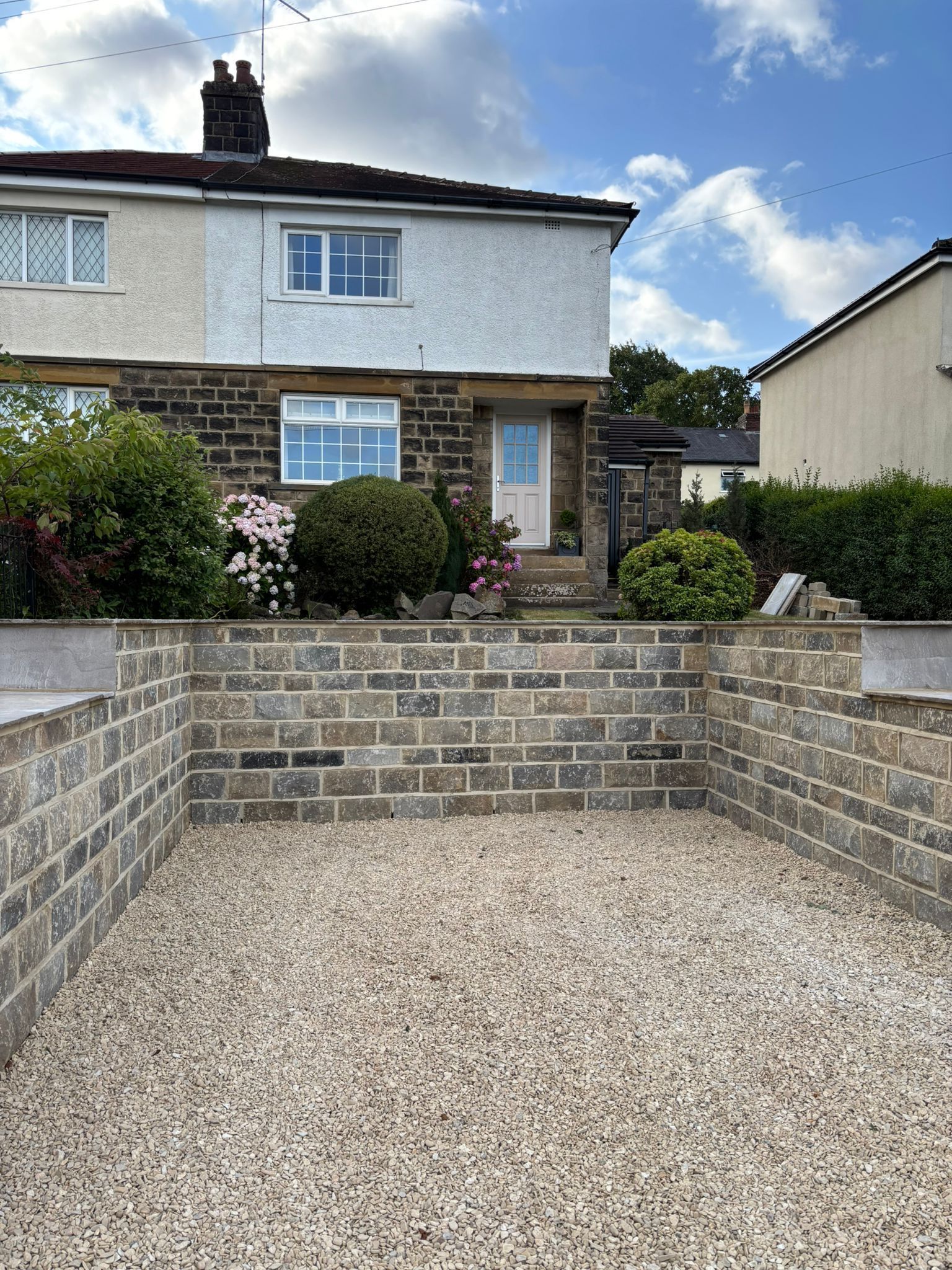 Small stone-front house with gravel driveway and low brick garden walls under a partly cloudy sky