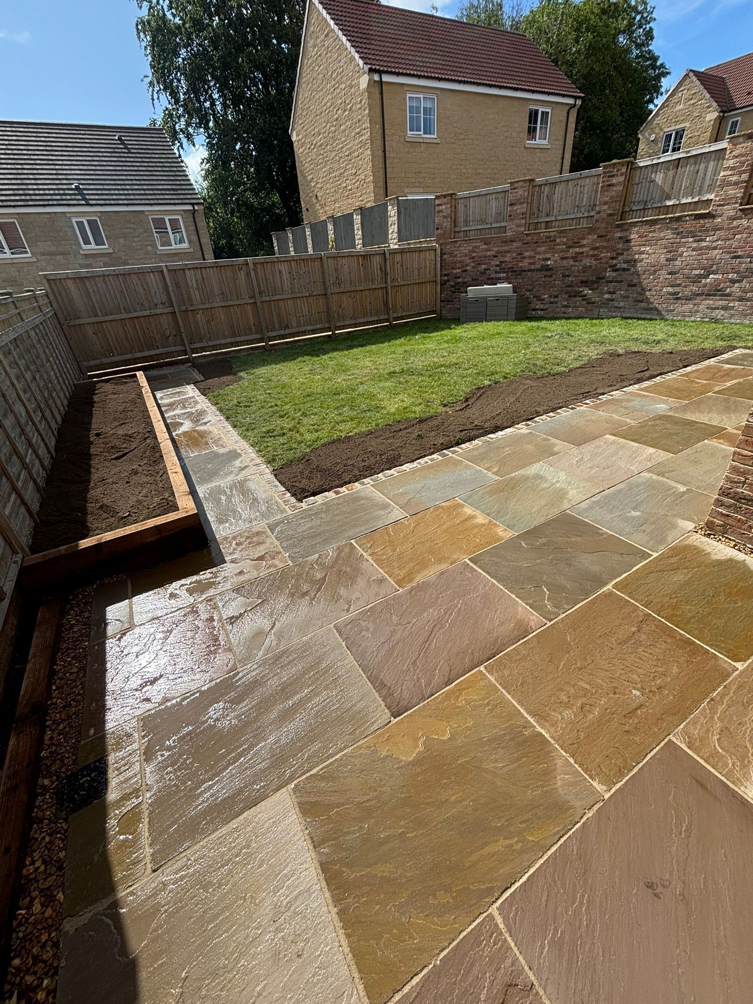 Backyard with wet stone patio, narrow grass strip, and wooden fence under bright sun