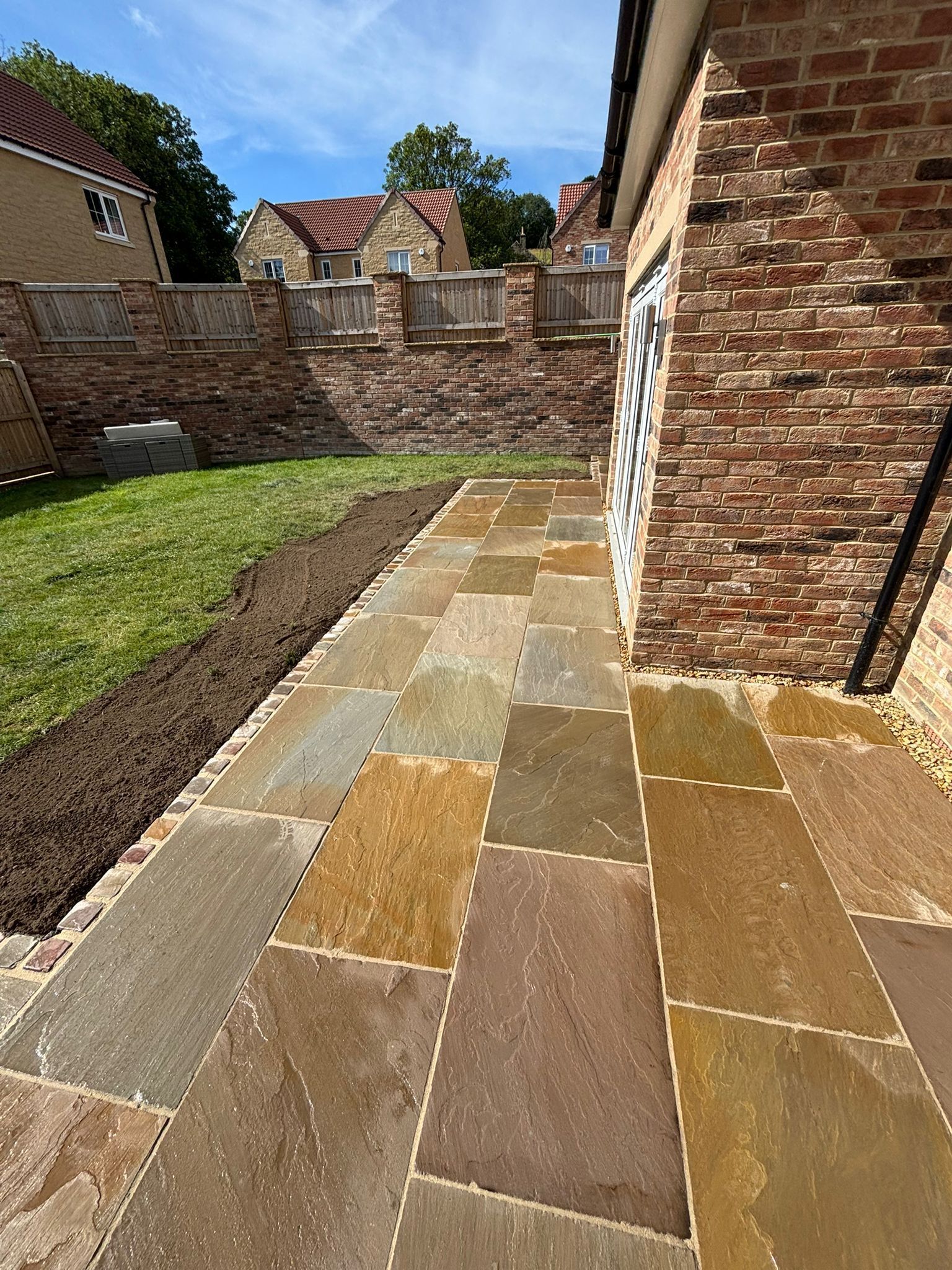Stone patio beside a brick house, with a narrow grass strip and garden wall in bright sunlight.