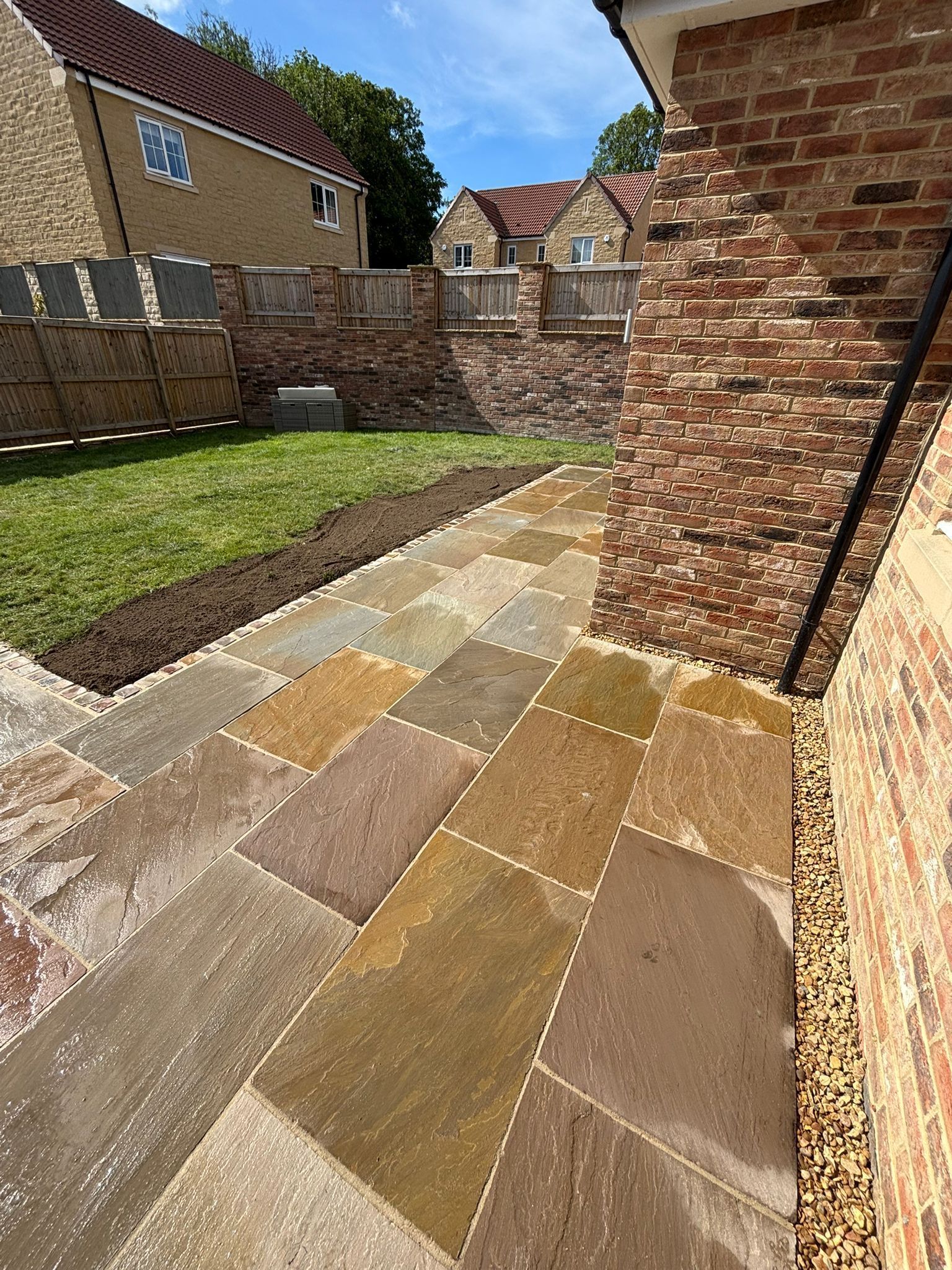 Stone-paved garden path beside a brick house and lawn in bright sunlight.