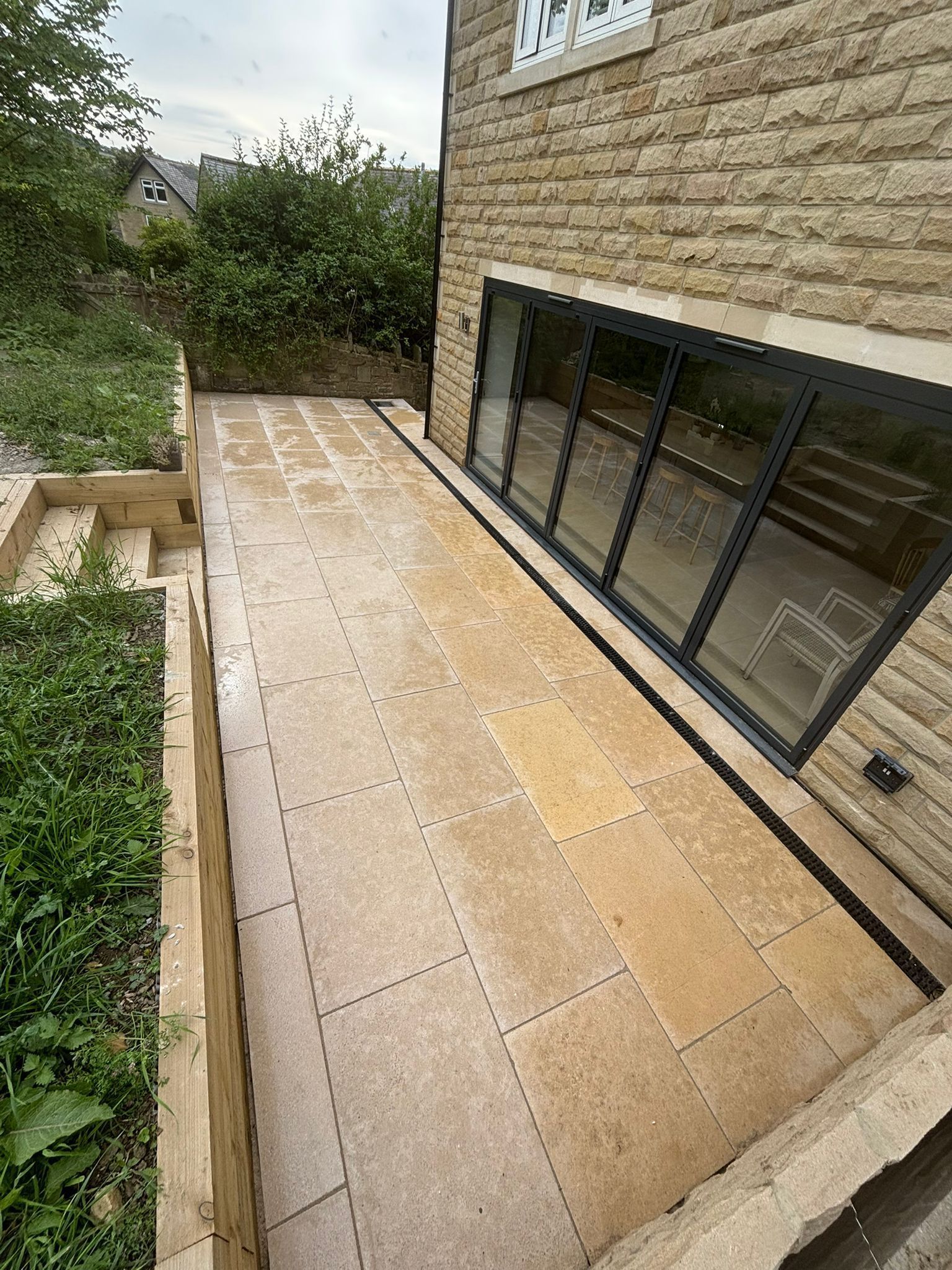 Stone patio beside brick house with large windows, bordered by greenery and a garden path