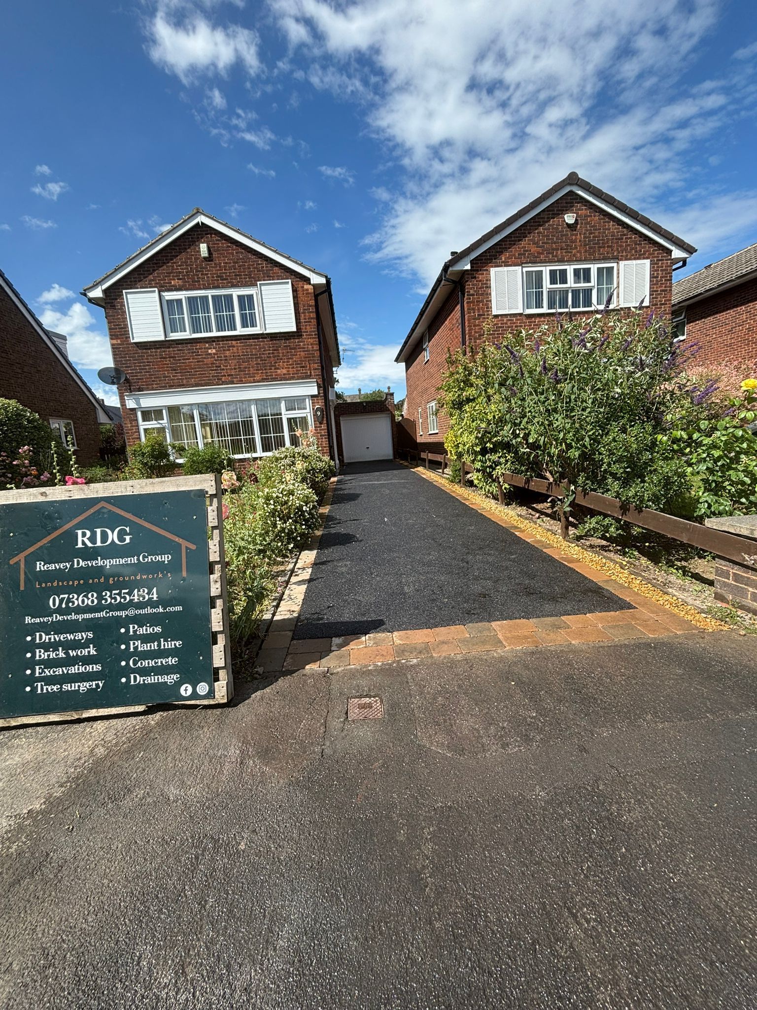 Two brick houses with a gravel driveway and a green roadside sign under a bright blue sky.