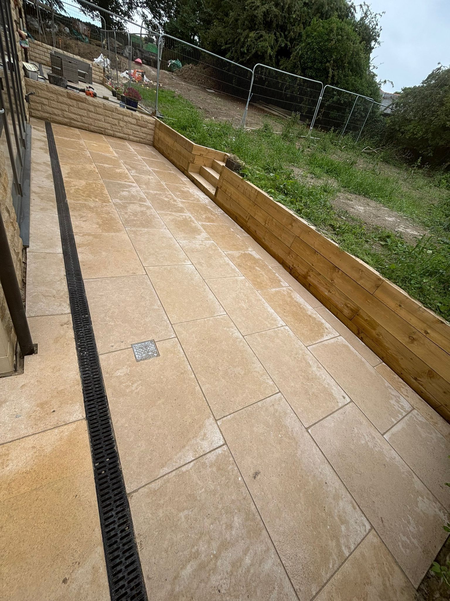 Stone patio path beside a grassy hillside, bordered by a low timber retaining wall and wire fence.
