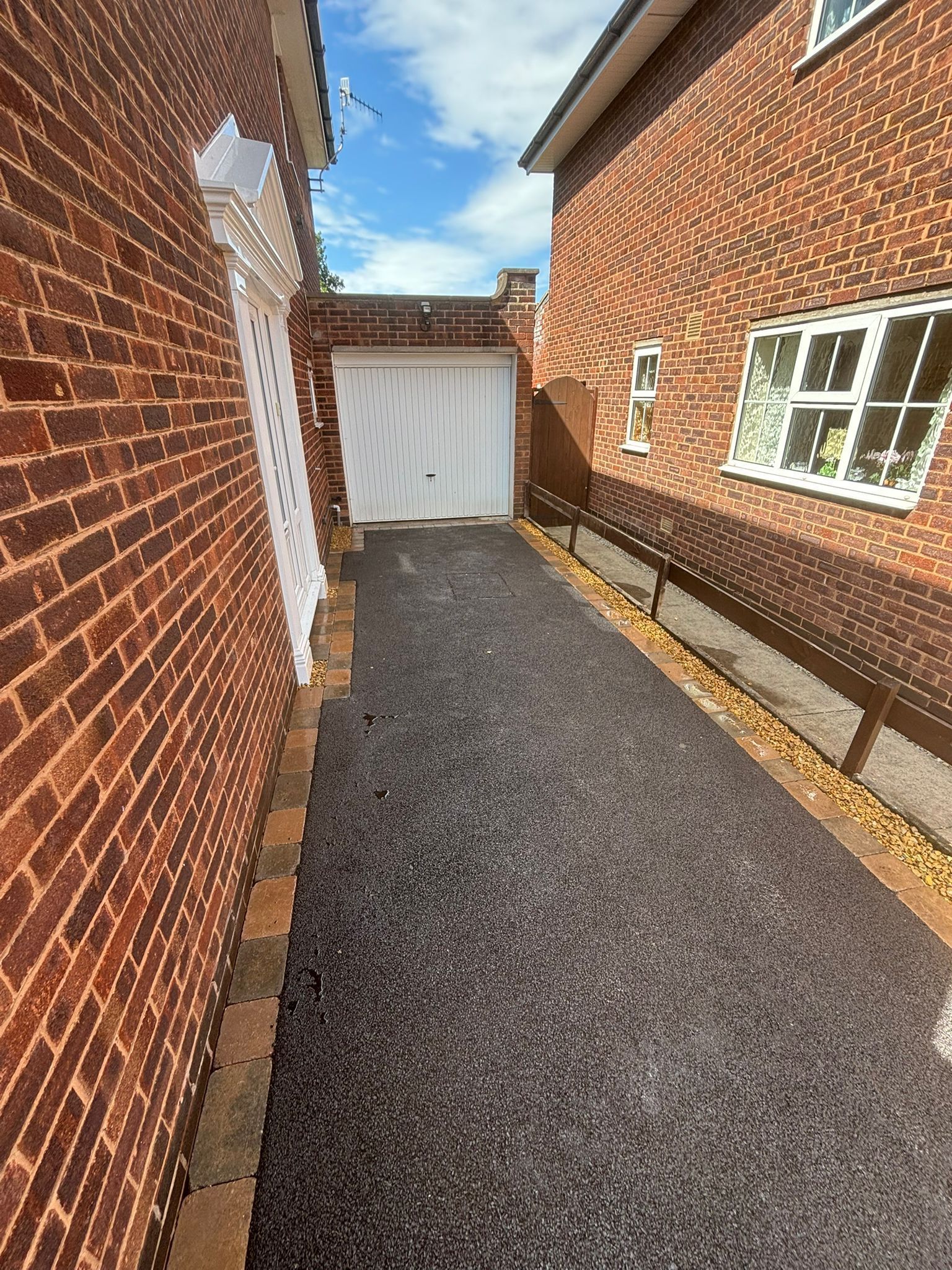 Narrow gravel driveway between red brick walls leading to a white garage door under a blue sky.