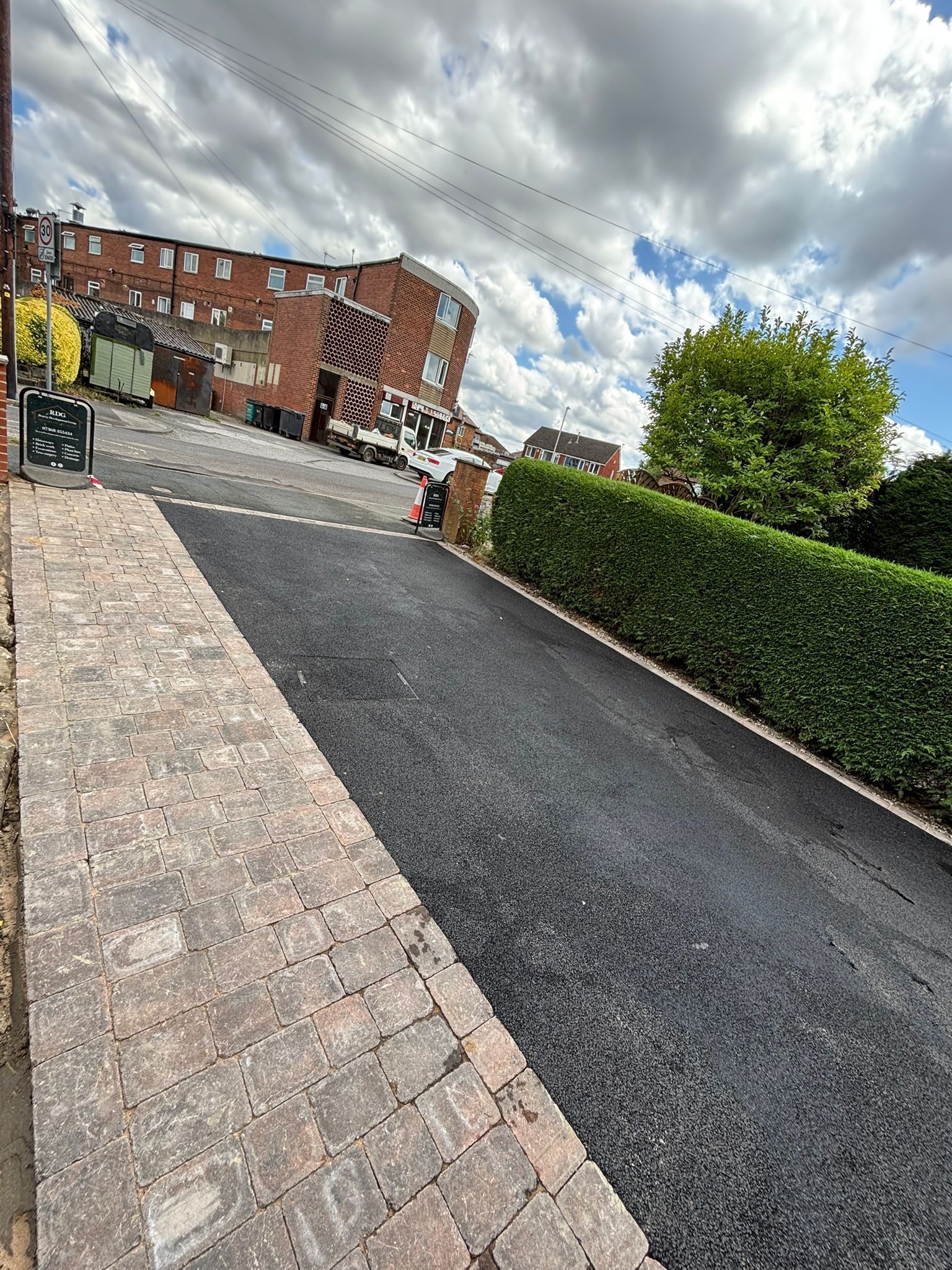 Sloped paved driveway beside trimmed hedge, leading to brick apartment buildings under cloudy sky.