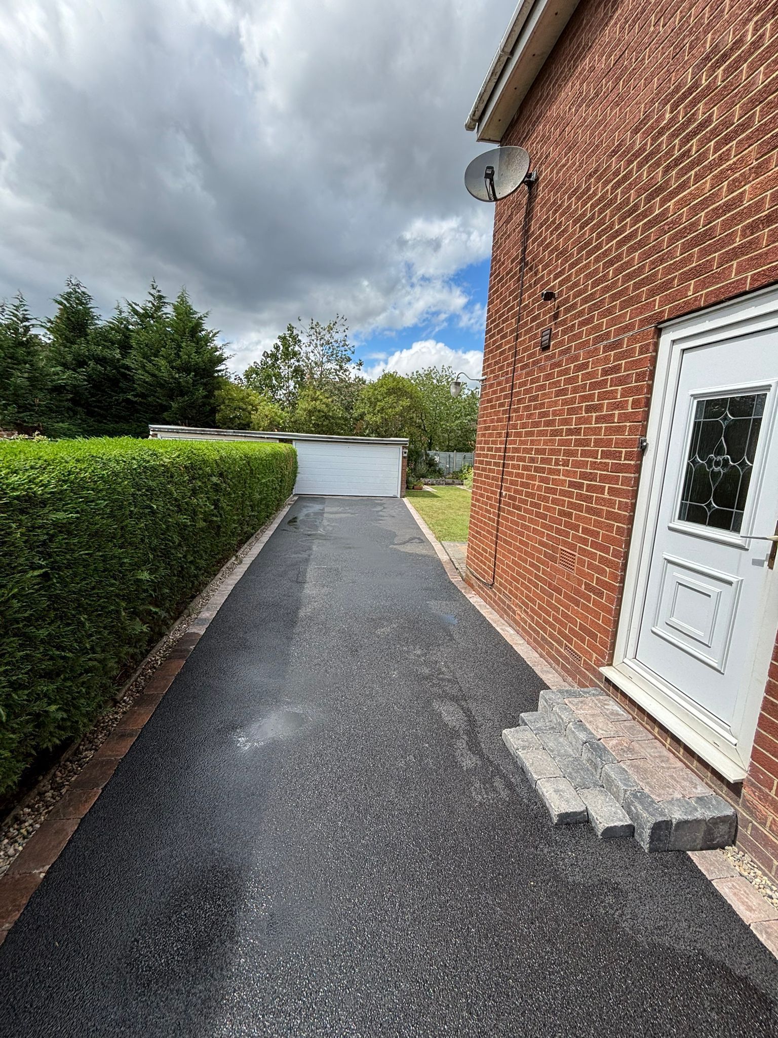 Gravel path beside a brick building with a white door, leading past a hedge toward trees and blue sky