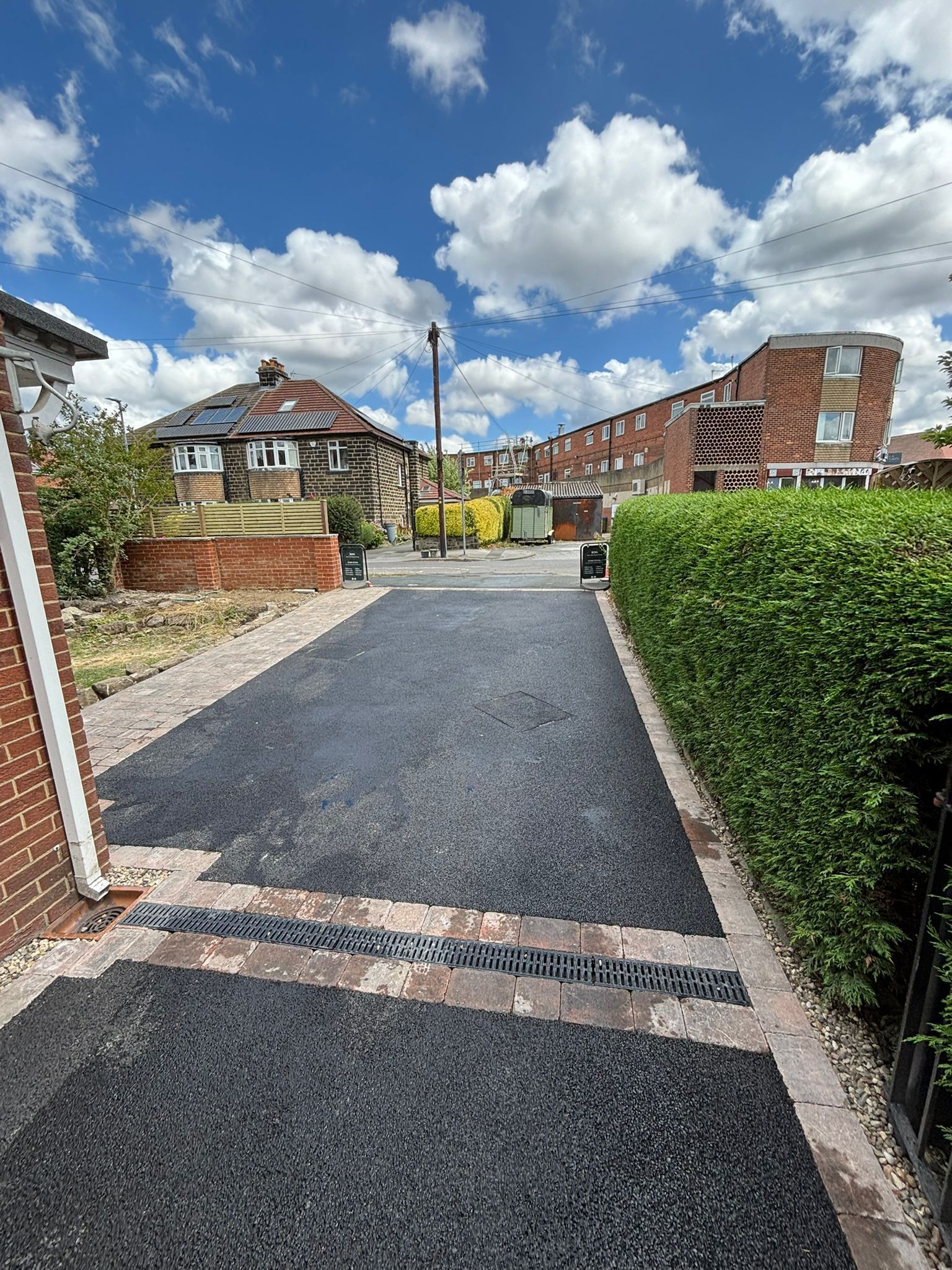 Gravel driveway between brick homes, with a hedge on the right and a blue sky with clouds overhead.