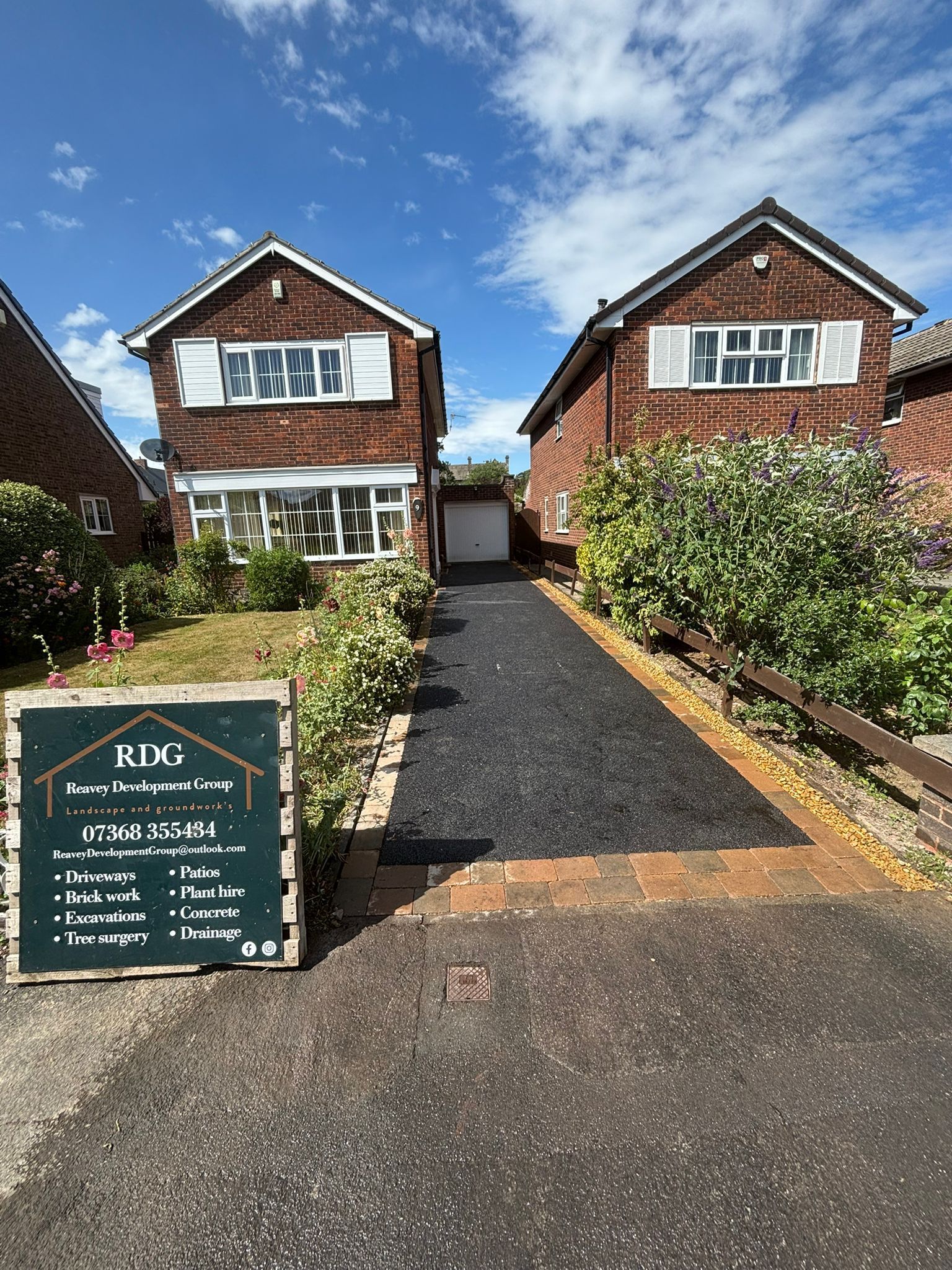 Two brick houses with a gravel driveway, front garden, and a signboard under a blue sky.