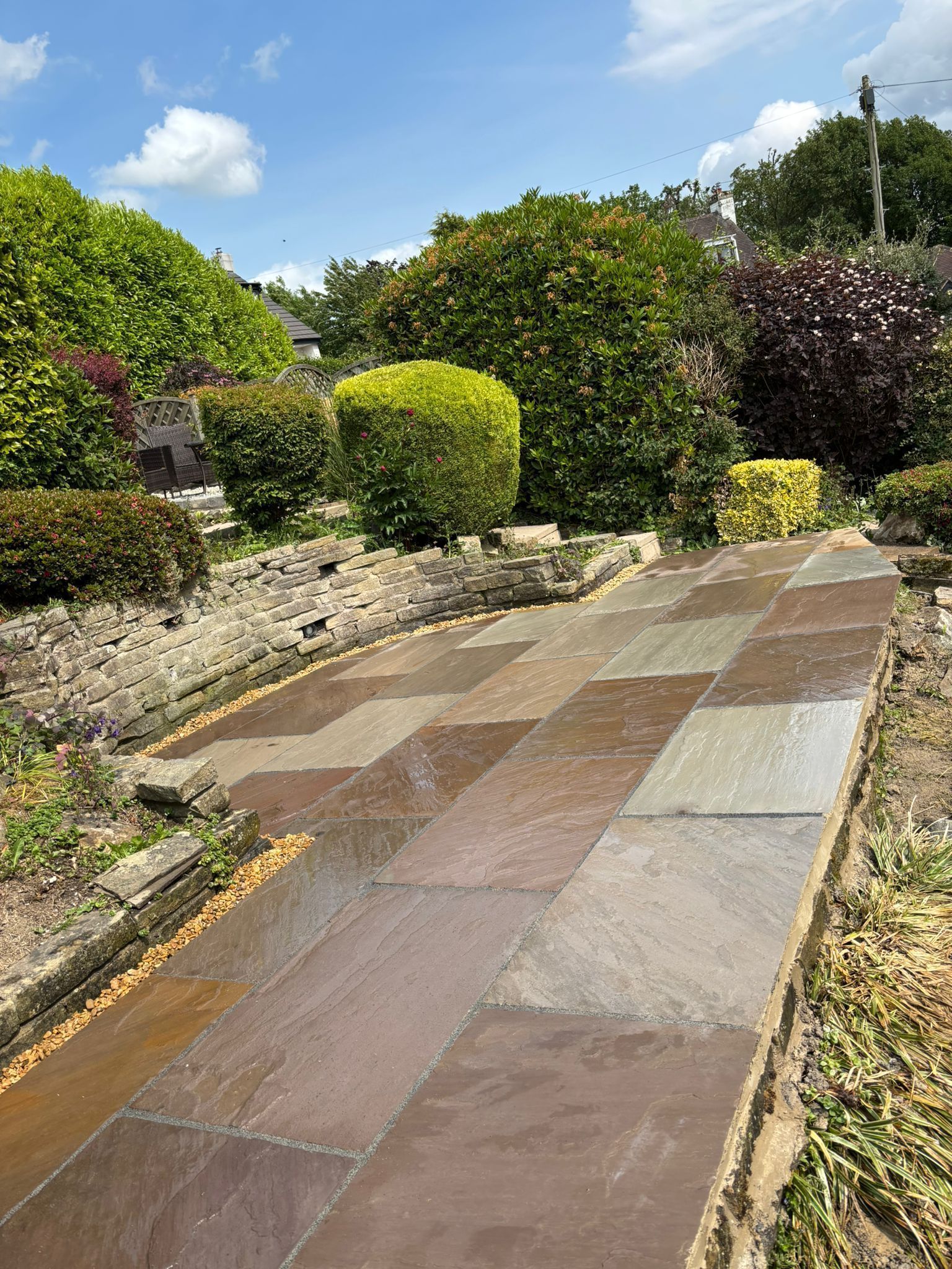 Wet stone garden path beside trimmed shrubs and trees under a bright sky
