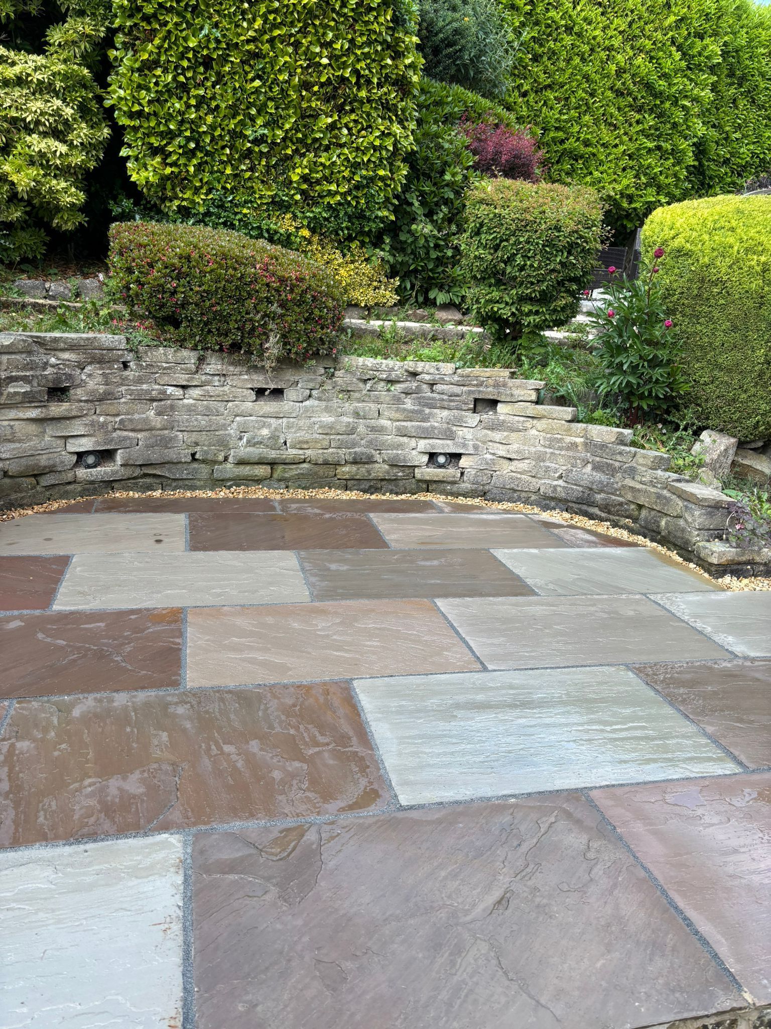 Paved patio with stone steps, low retaining wall, and manicured green shrubs in a garden.