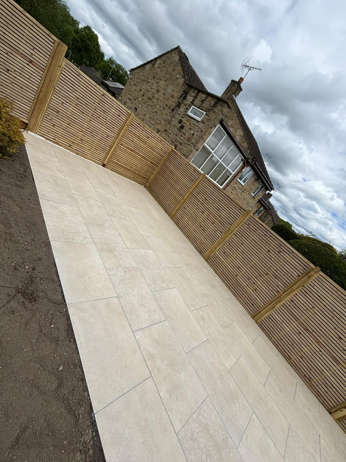 Stone house with a tiled roof beside a pale paved patio and brick wall under a cloudy sky