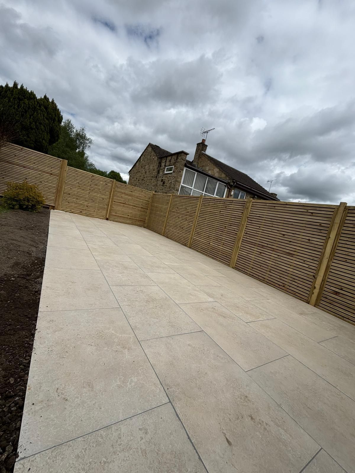 Large paved patio with wooden fencing, trees, and a cloudy sky behind a house