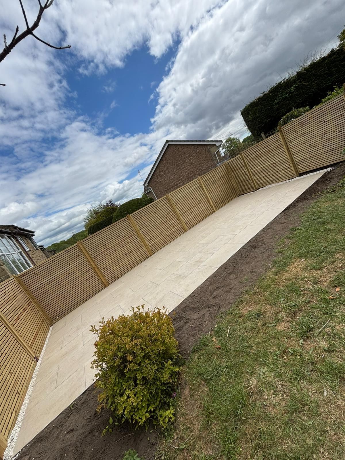 Tilted view of a fenced backyard with a paved patio, small bush, and cloudy sky