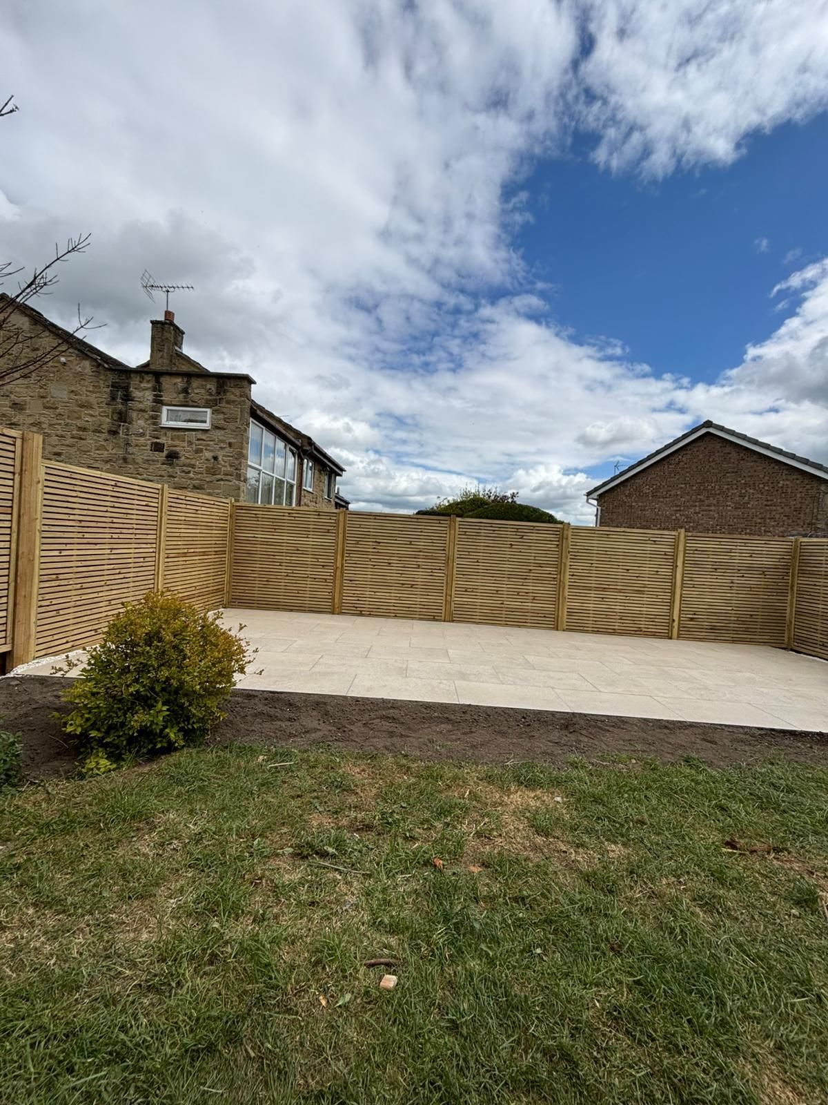 Backyard with grassy slope, cream fence, brick wall, and cloudy sky behind a house