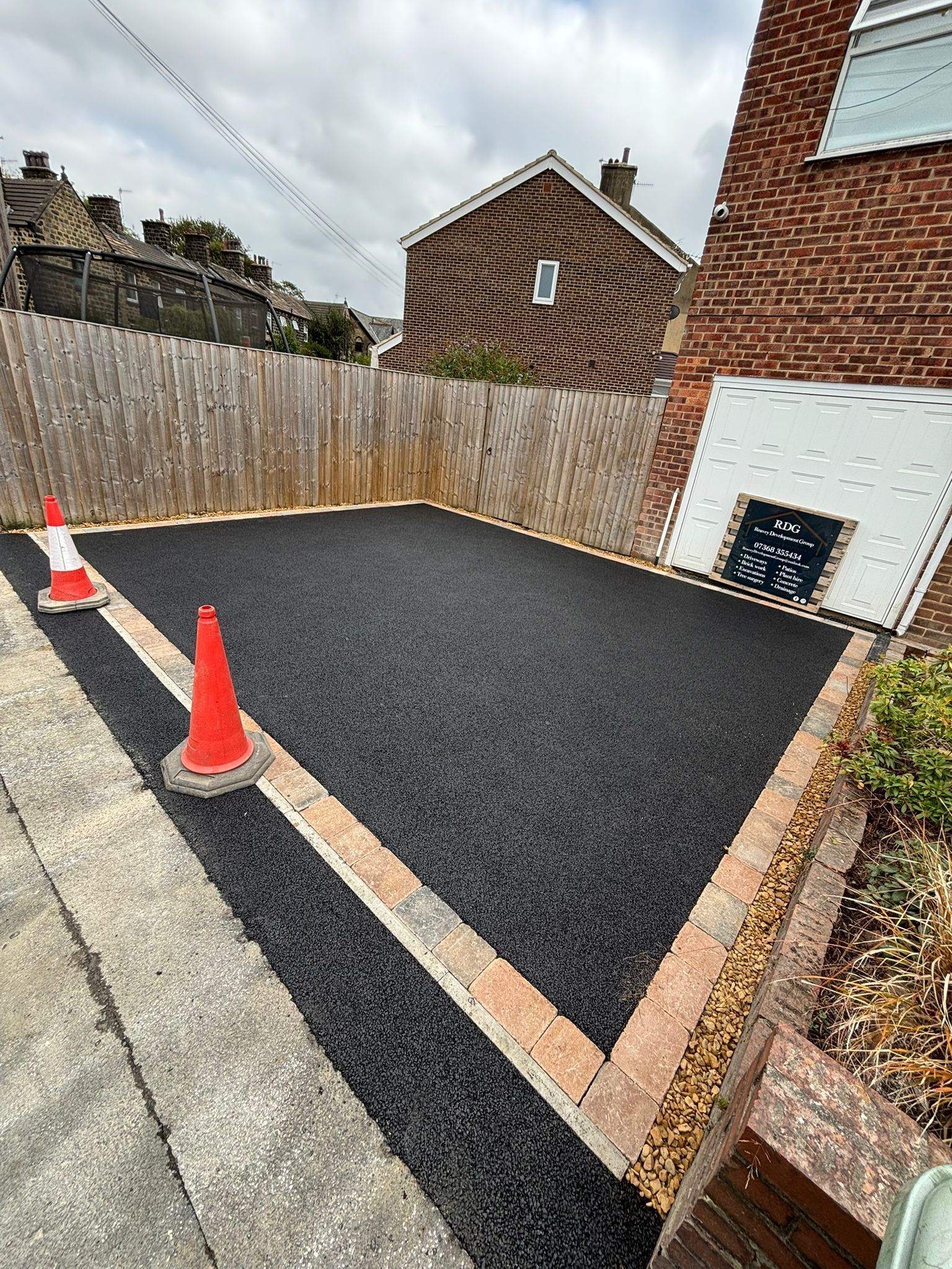 Freshly paved black driveway with two orange cones beside a brick house and wooden fence