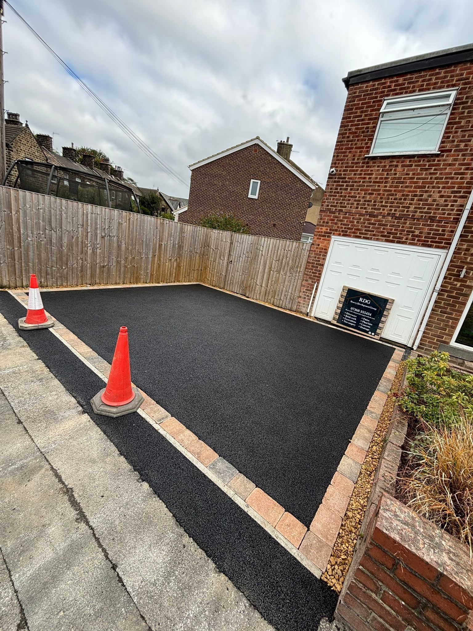 Freshly paved driveway with orange cones beside a brick house and wooden fence