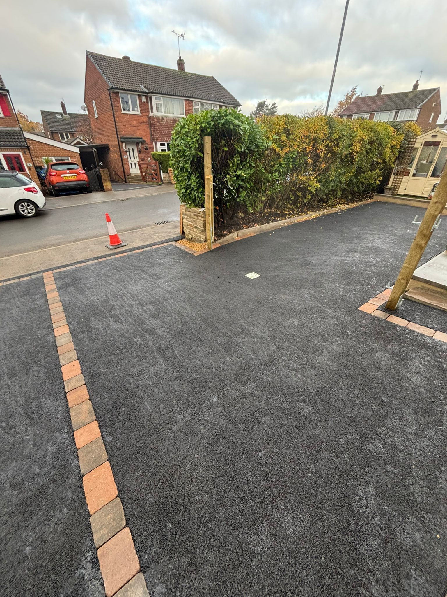 Newly resurfaced driveway beside brick houses, with hedge, stone pillars, and traffic cones by the street