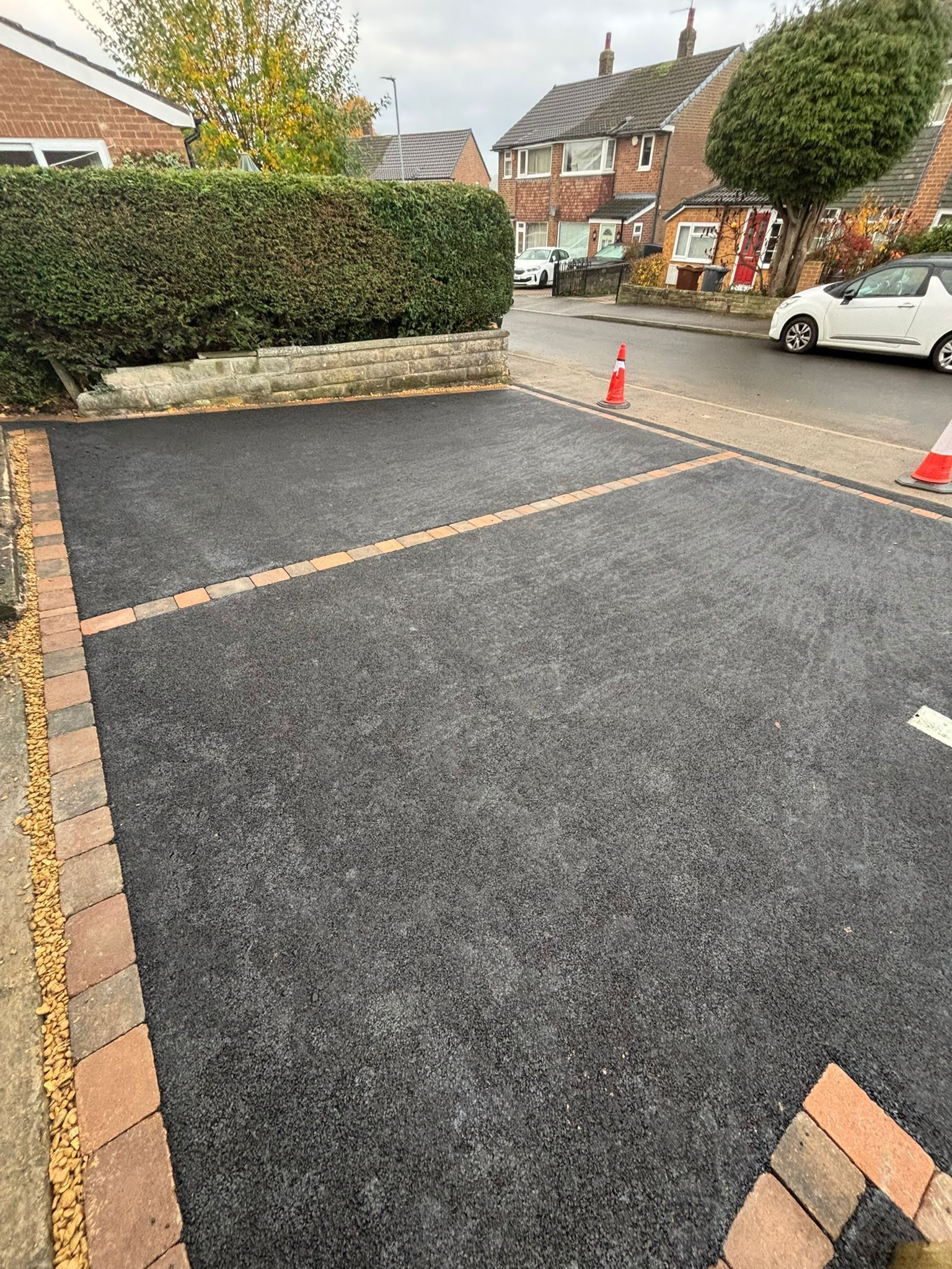 Freshly resurfaced parking area with orange barrier tape and traffic cone beside a hedge and brick houses