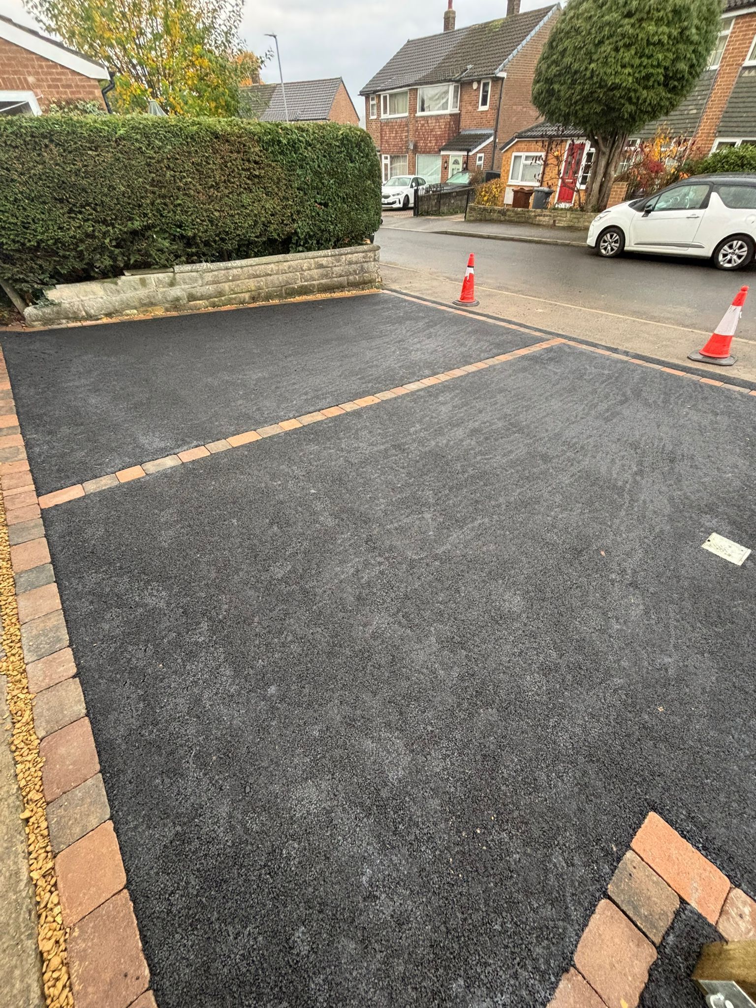 Empty asphalt parking bay bordered by brick pavers, with traffic cones and houses in the background