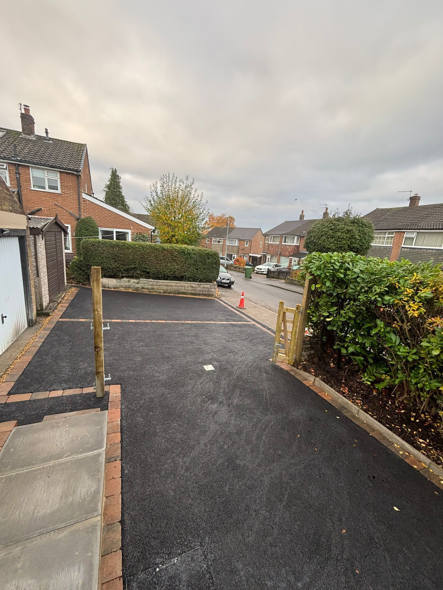 Residential street view with a black paved driveway, hedges, brick houses, and cloudy sky.