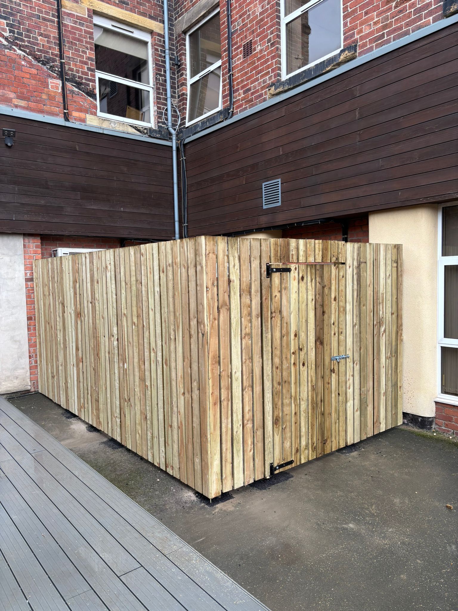 Wooden dumpster enclosure beside a brick building on a paved courtyard